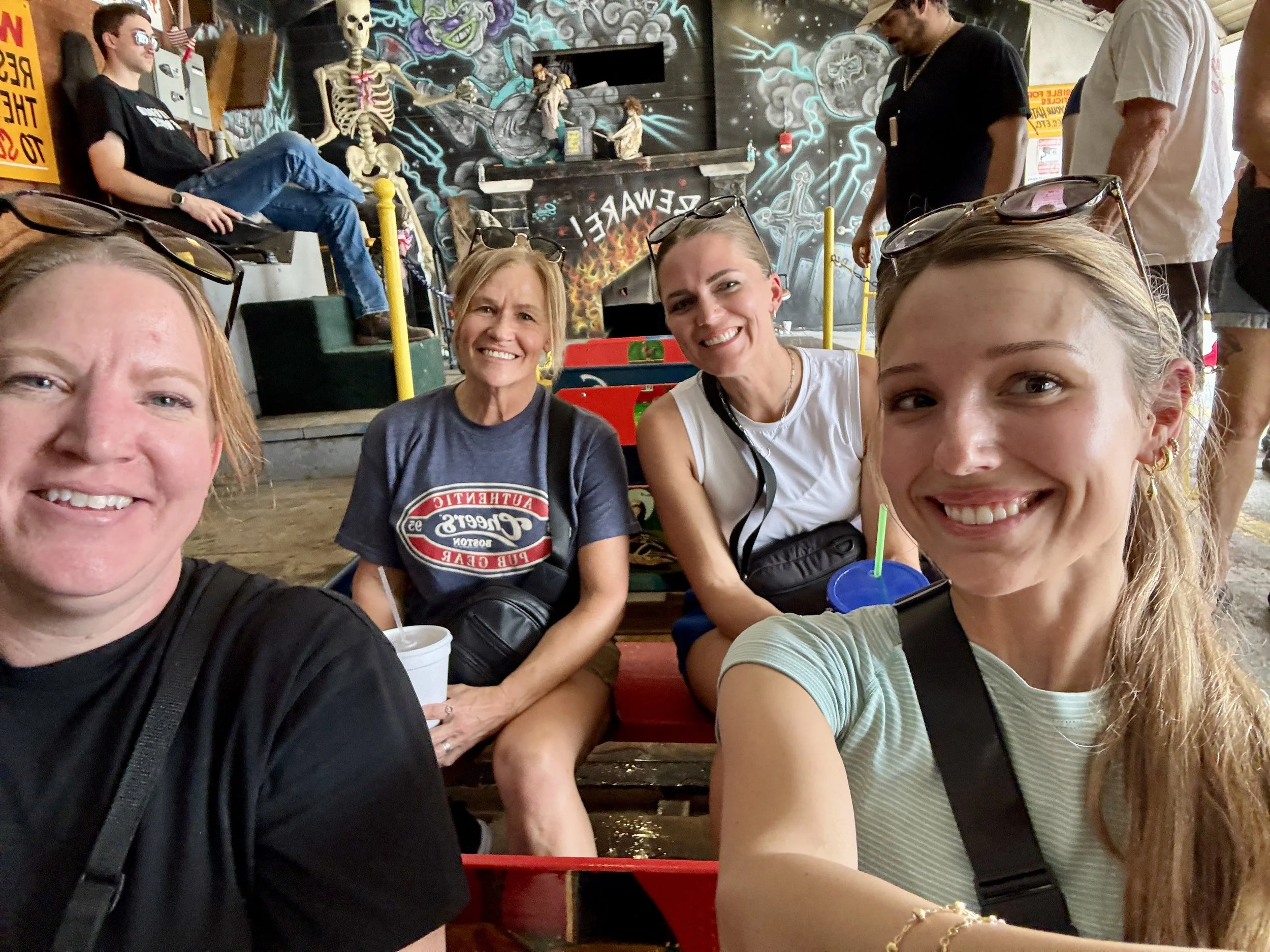 A group of smiling women and young girls taking a selfie at an indoor amusement or arcade area. The background features colorful graffiti-style artwork with skulls, a fireplace, and the word 'DEWALT'. Other people are visible in the background, some 