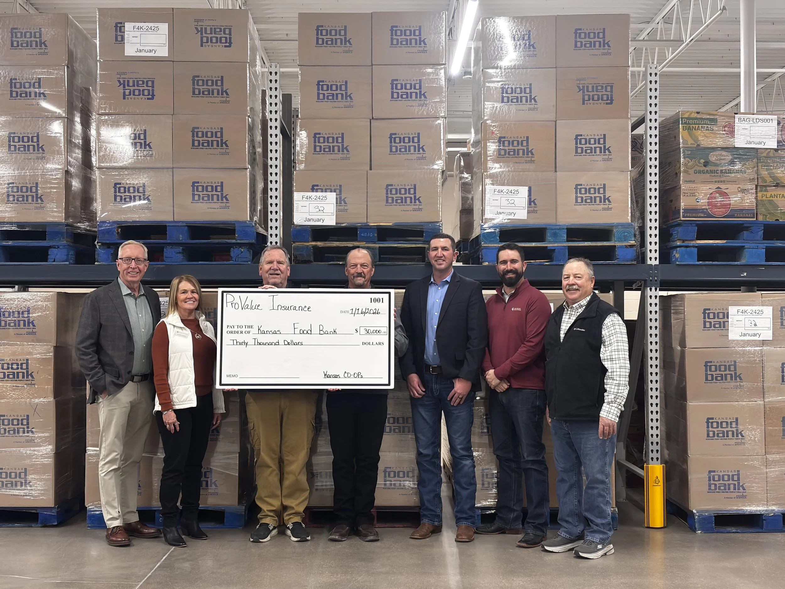 Group of seven people standing inside a warehouse in front of stacks of boxes labeled Kansas Food Bank, holding a large check for $30,000 made out to Kansas Food Bank.