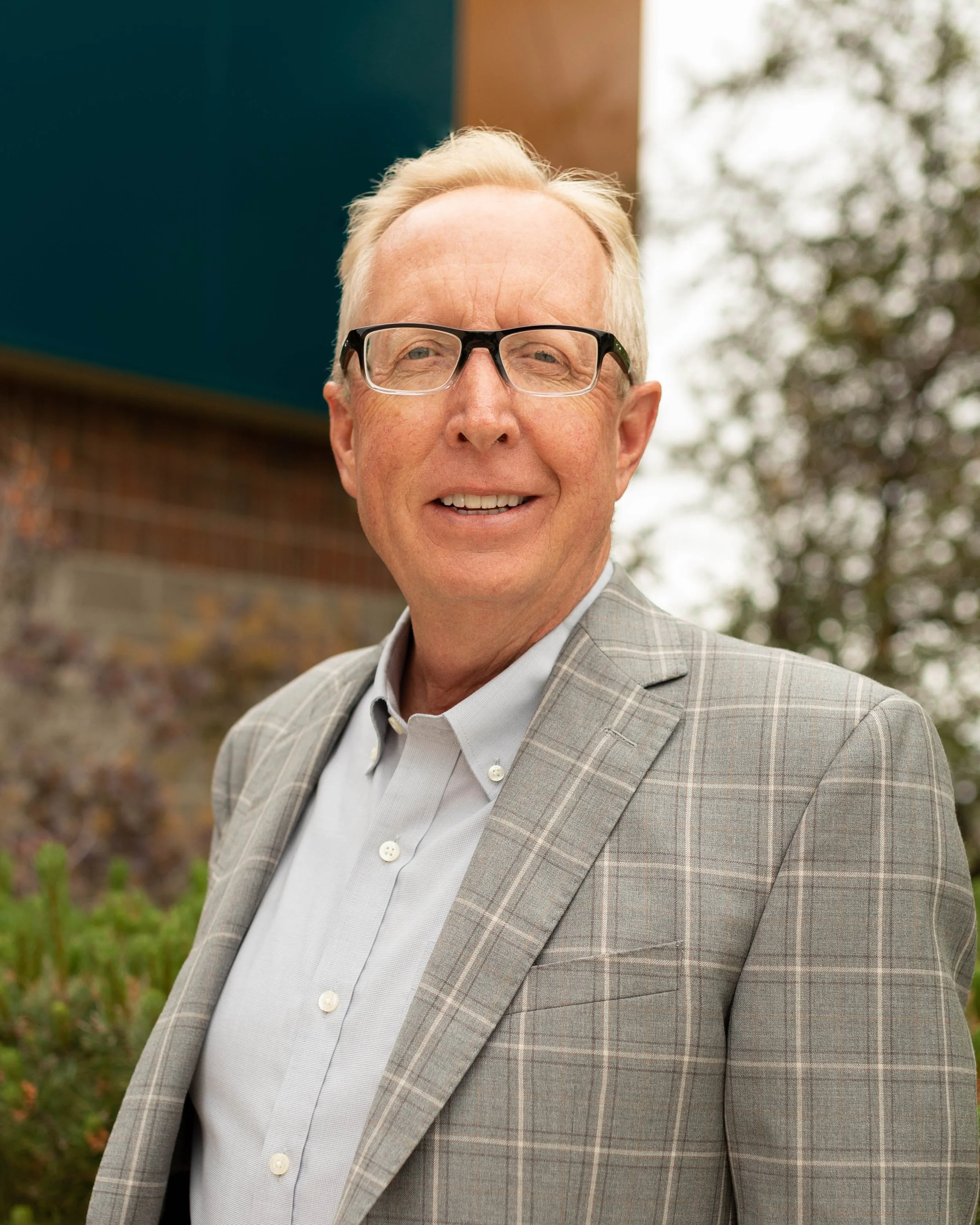 Close-up of a smiling middle-aged man with glasses, wearing a gray checkered blazer and a white shirt, standing outdoors with blurred trees and a building in the background.