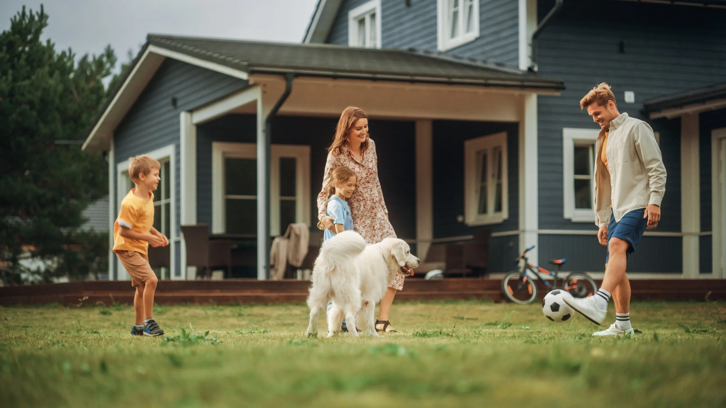 A family playing soccer outside their house, with a dog and bicycles nearby.