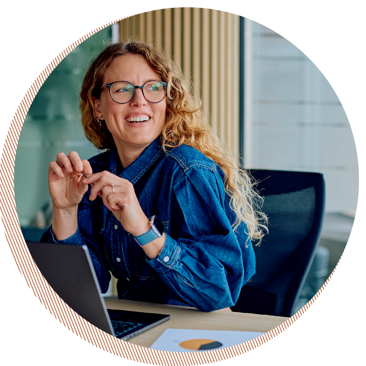 A woman with curly red hair and glasses smiling while sitting at a desk with a laptop and papers, in an office setting.
