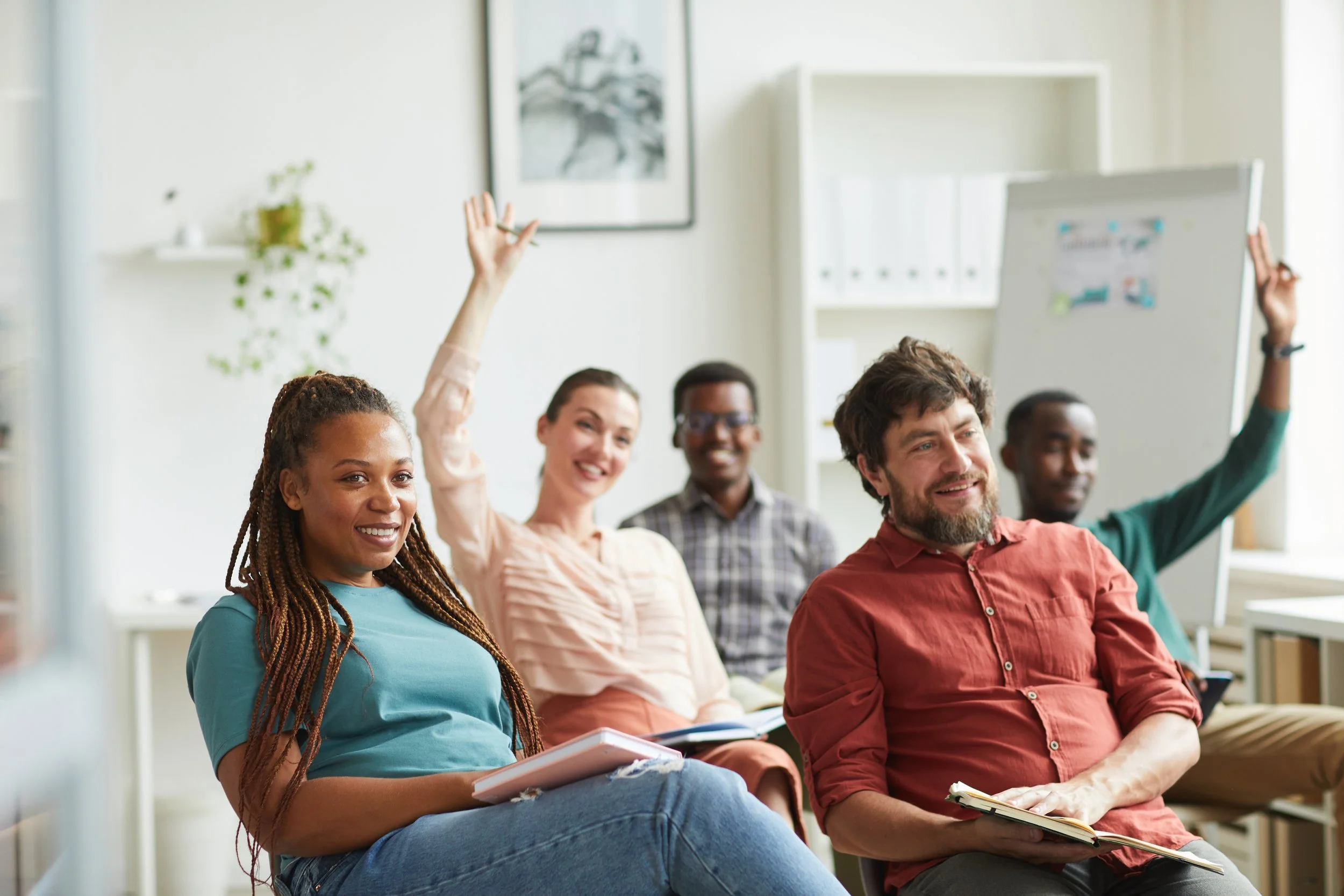 A diverse group of people sitting in a classroom or meeting room, some raising their hands, others smiling and holding notebooks.