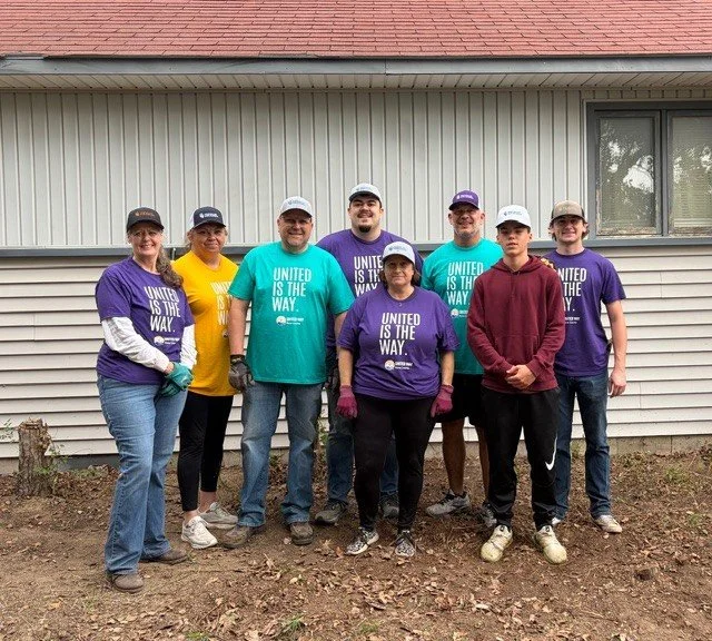 Group of eight volunteers, including both men and women, standing outdoors in front of a house, participating in a community cleanup or renovation effort. They are wearing casual clothing and some are wearing gloves and hats.