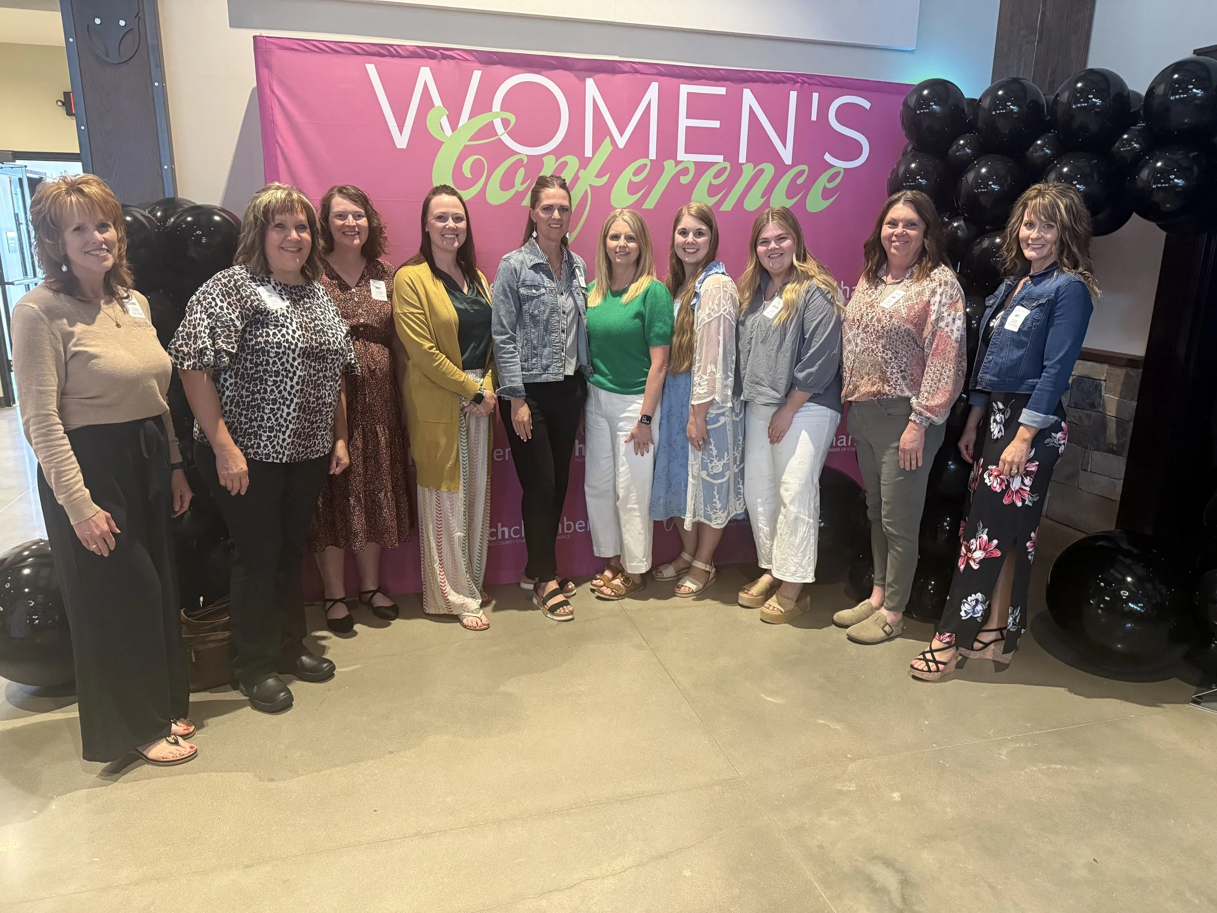 A group of women standing together in front of a pink Women's Conference backdrop with black balloons on either side, inside a building.