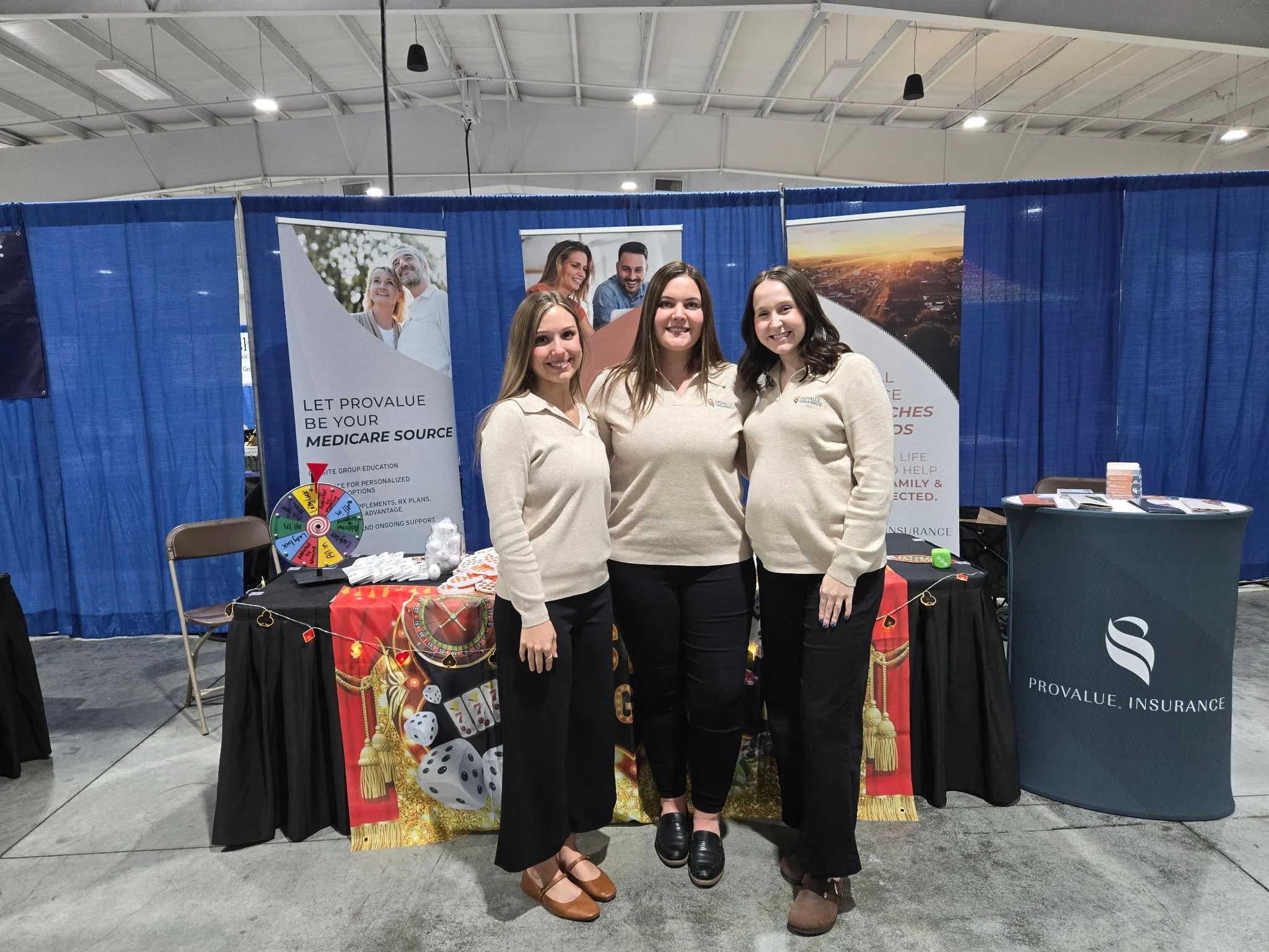 Three women standing in front of a booth at an indoor event. The booth has blue curtains and promotional banners, with one banner displaying images of people and text about Medicare insurance. There is a table with marketing materials and a decorativ