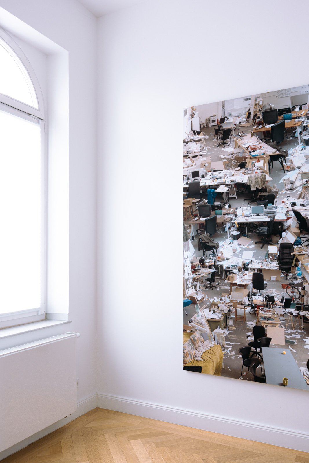Ein sauberer Raum mit einem großen Fenster auf der linken Seite und einem Bild an der rechten Wand. Das Bild zeigt einen unordentlichen Arbeitsplatz mit vielen Papieren und Bürostühlen. Der Raum hat einen hölzernen Parkettboden und weiße Wände.