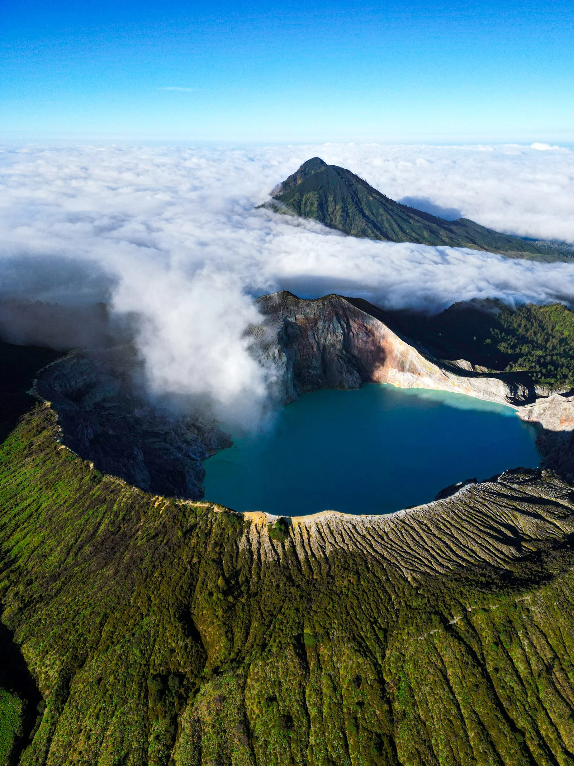 A volcanic crater lake surrounded by green slopes with a mountain and clouds in the background.