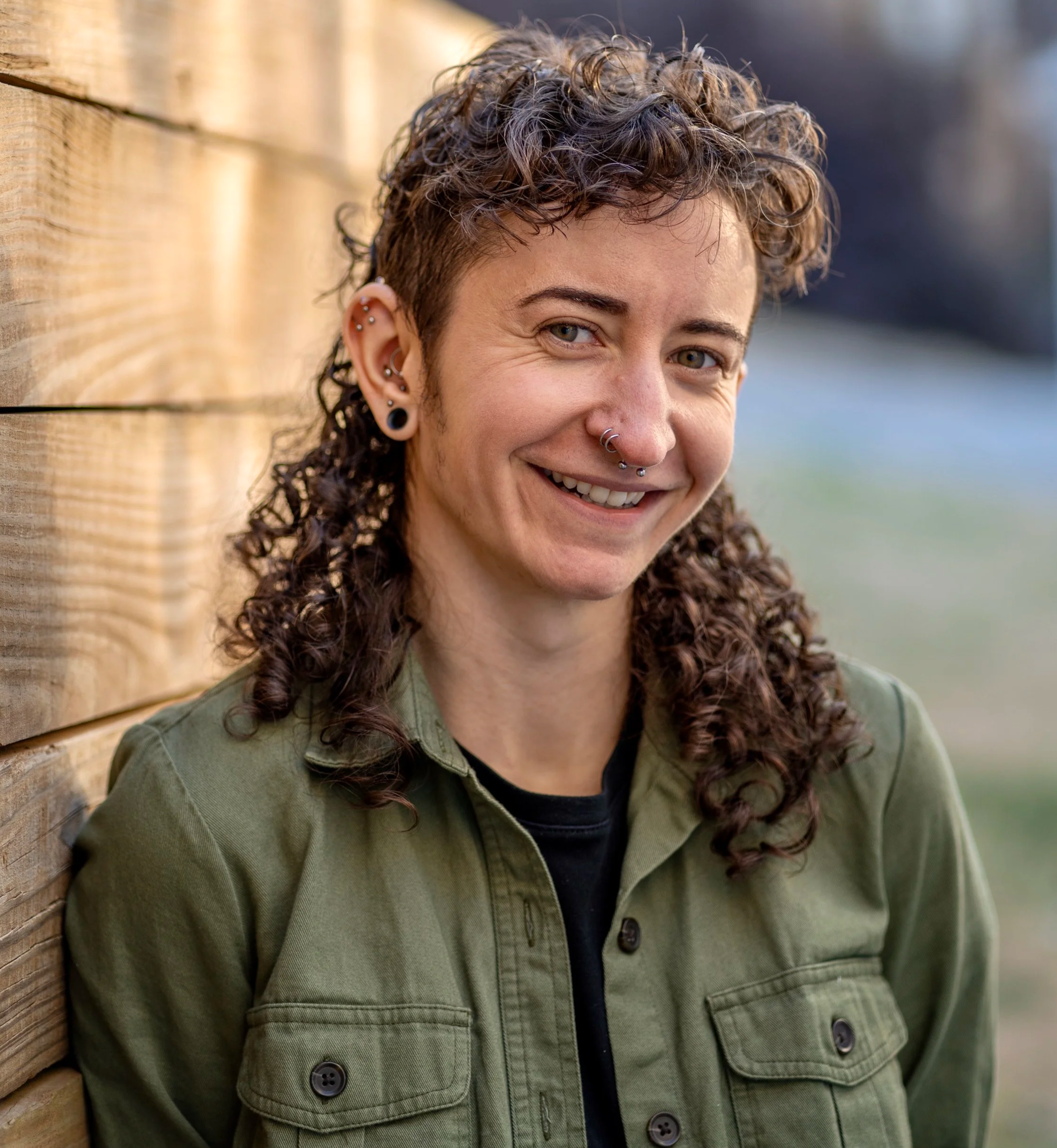 A non-binary trans person with curly brown hair, multiple earrings, and a septum piercing smiling, wearing an olive green jacket, leaning against a wooden fence outdoors.