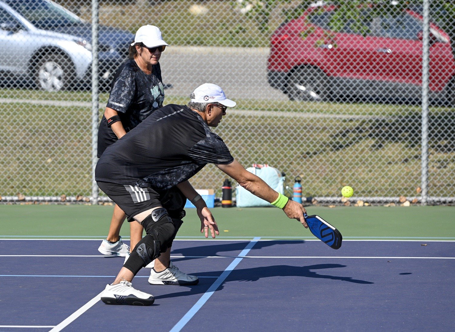 Two older people playing pickleball on a court, one preparing to hit a yellow ball with a paddle, with solitude behind a chain-link fence.