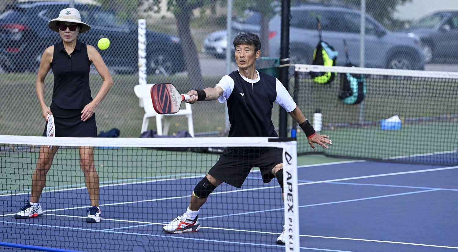 Two elderly people playing pickleball on an outdoor court, one is hitting a ball with a paddle while the other watches nearby.