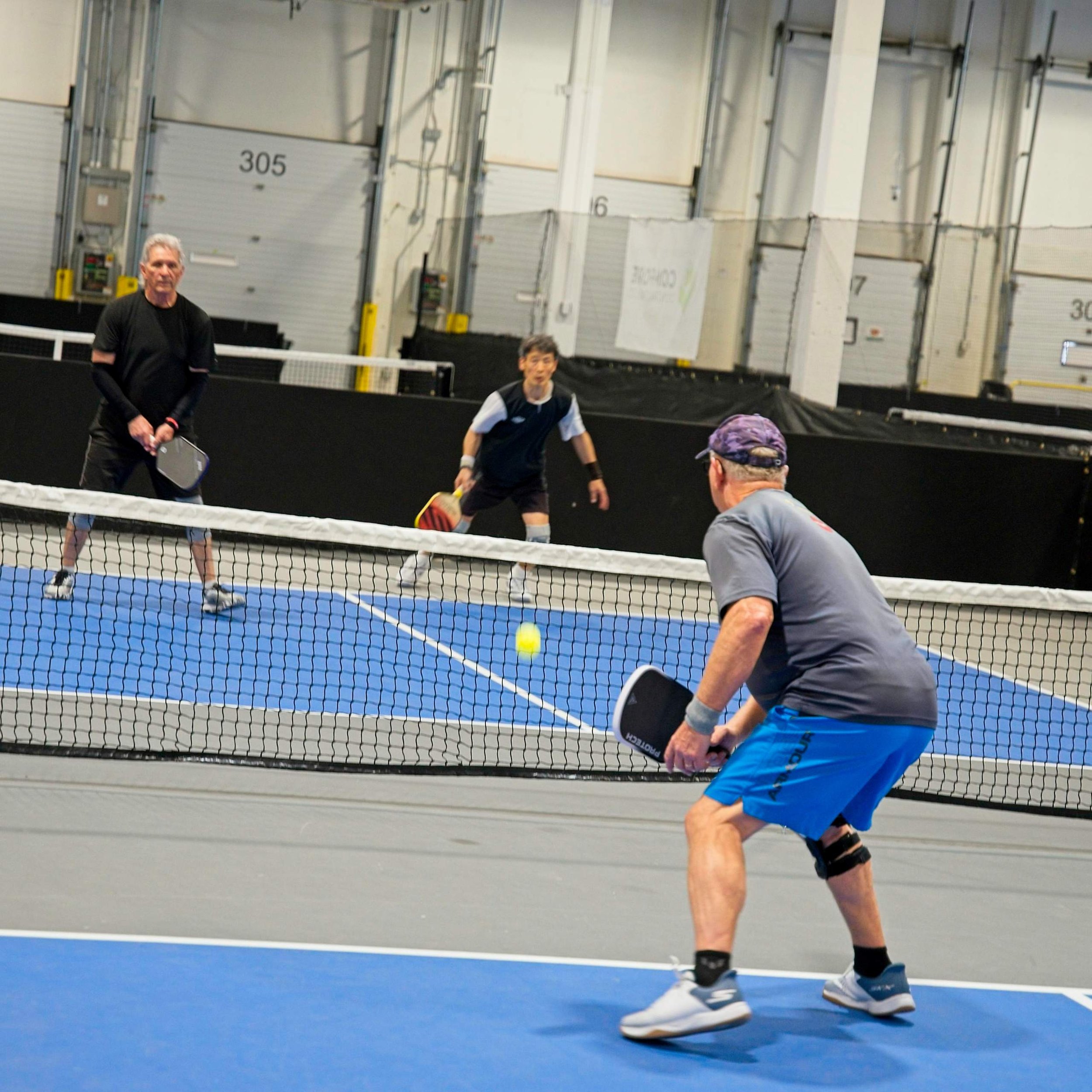 Two men playing pickleball, one is standing with a paddle in hand and the other is preparing to hit the ball, on an indoor court with a net and numbered walls.