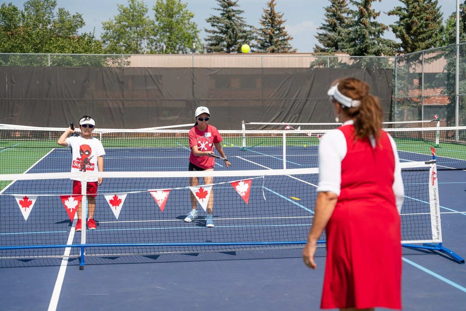 A woman in a red dress and white top playing pickleball on an outdoor court, with two young girls nearby, one wearing a white T-shirt and red shorts, the other wearing a red T-shirt and black shorts, both wearing sunglasses and visors, with a net, te