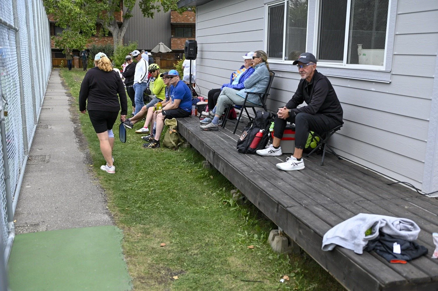 A group of tennis players and spectators sitting on a wooden deck outside a sports facility, with some holding tennis rackets and wearing athletic clothing, while others are seated or standing, waiting or resting.