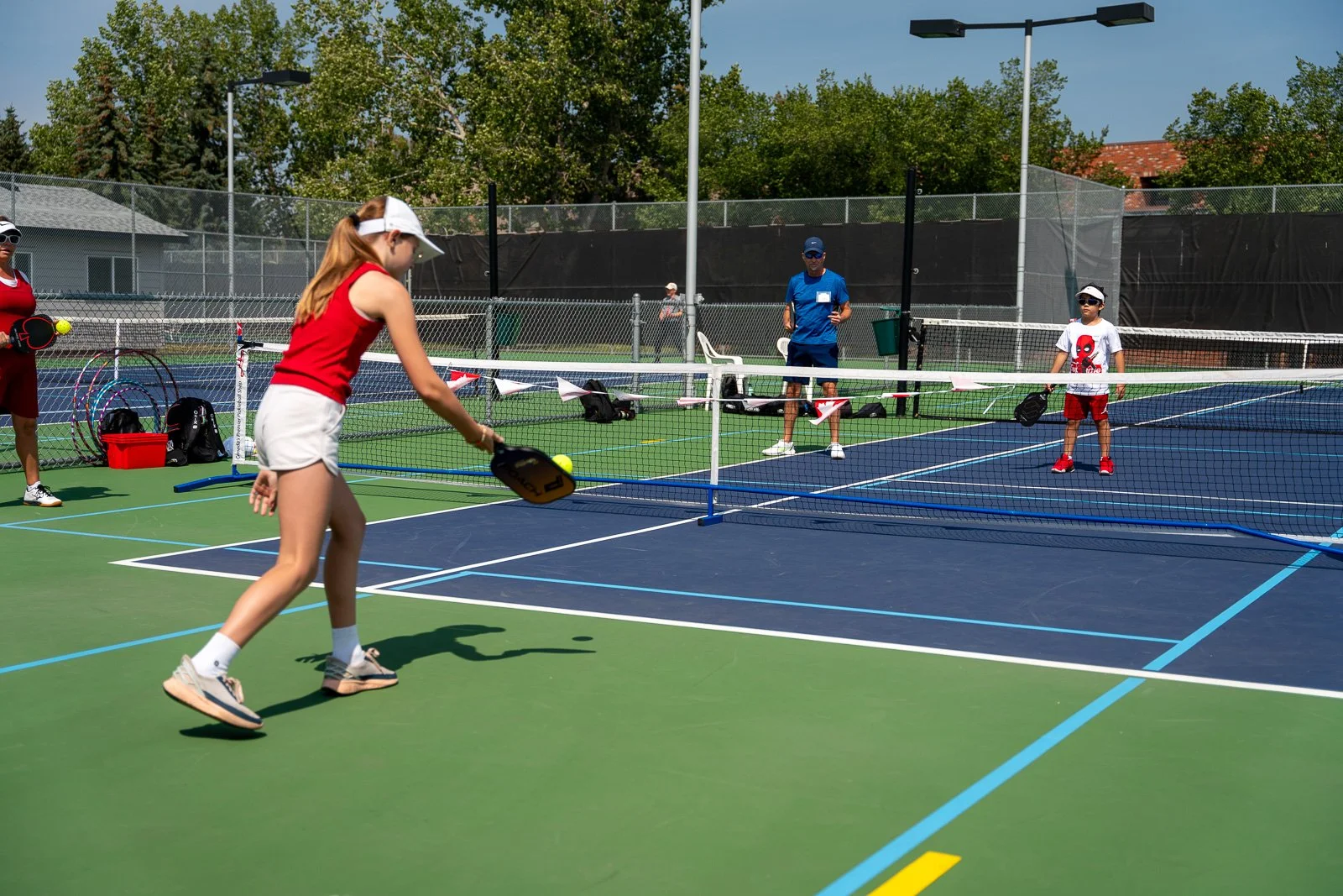 Kids playing pickleball on an outdoor court with net and officials watching, trees and buildings in the background under clear sky.