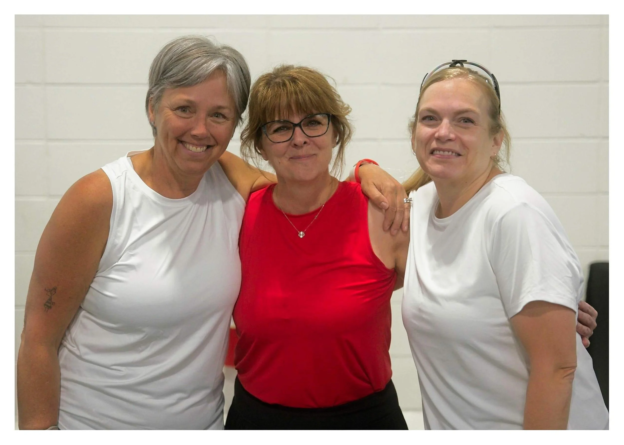 Three women smiling and standing close together with arms around each other against a plain white wall.