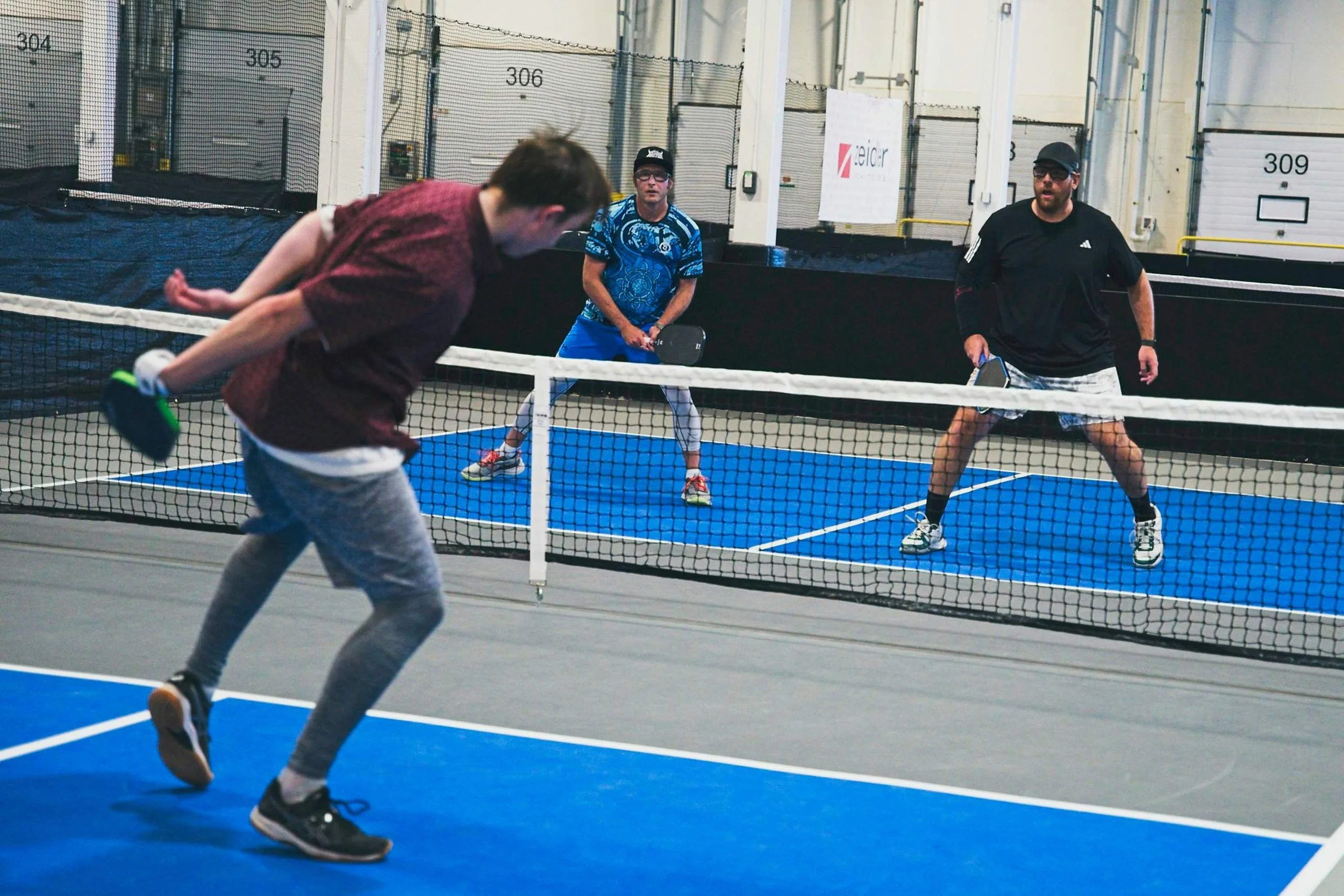 Two men playing pickleball with a woman in the background indoors on a blue court.