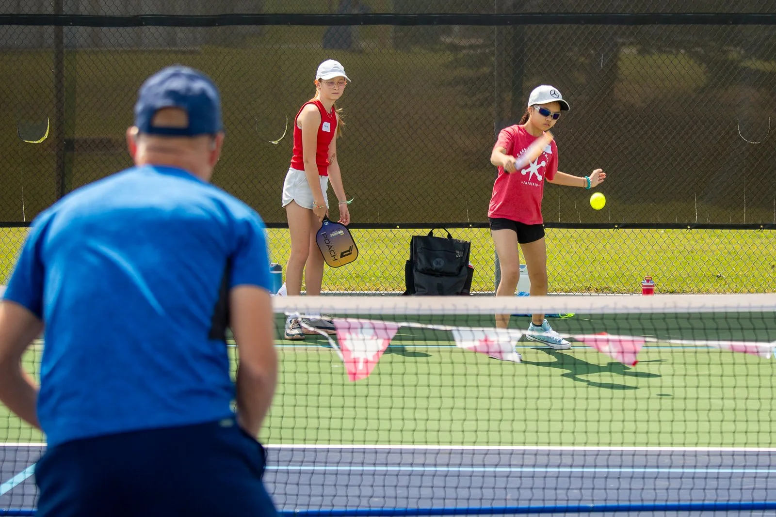 Two young girls playing pickleball on an outdoor court, with a man watching from the foreground. The girl on the right is hitting the ball, wearing sunglasses and a cap. The girl on the left is holding a paddle and observing. The court has a net with