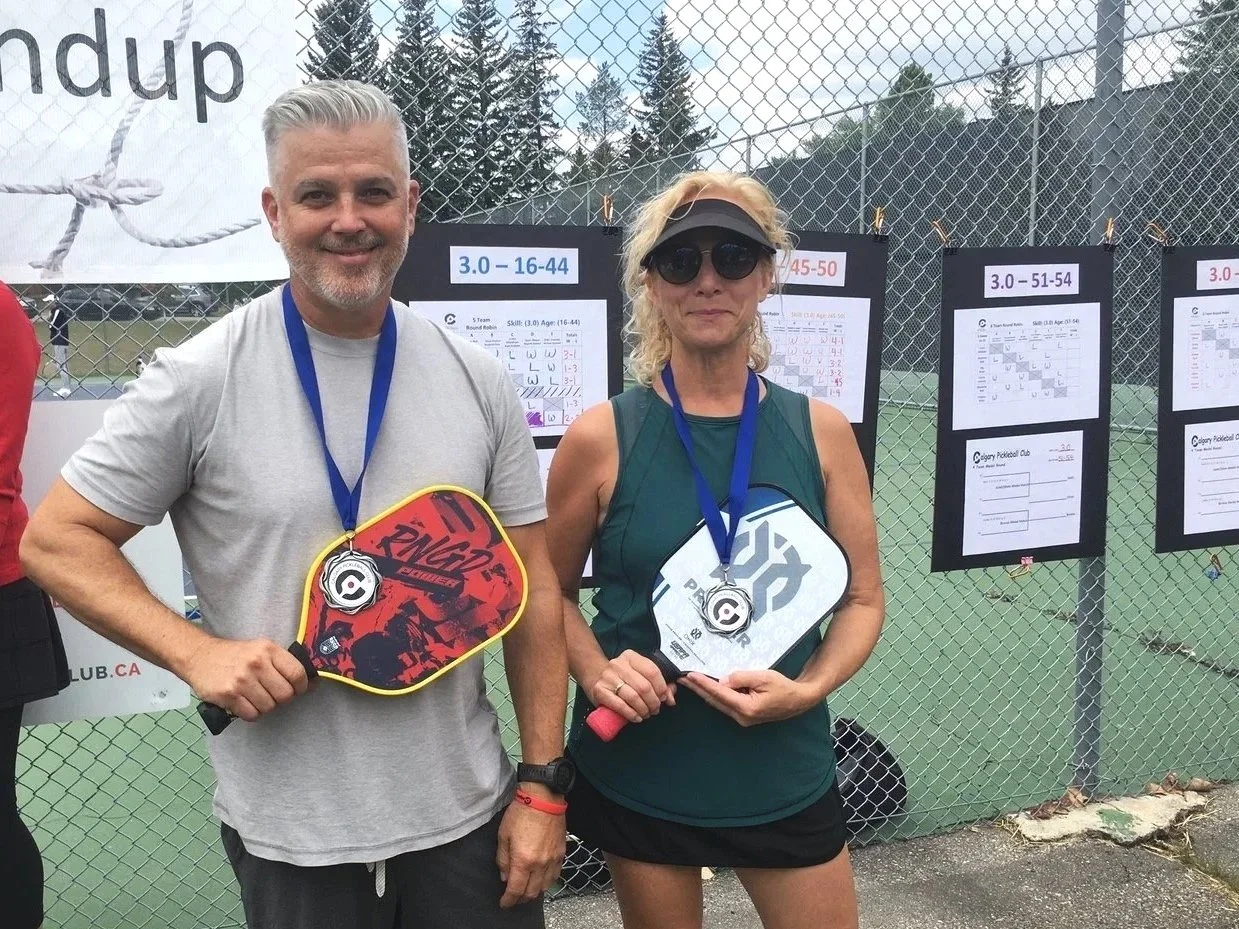 Two people holding pickleball paddles with medals around their necks, standing in front of a fence with score sheets and a banner in the background.