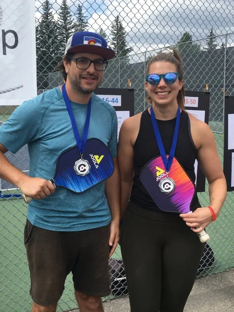 Two people standing outdoors near a tennis or pickleball court, wearing medals around their necks and holding paddles, smiling at the camera.