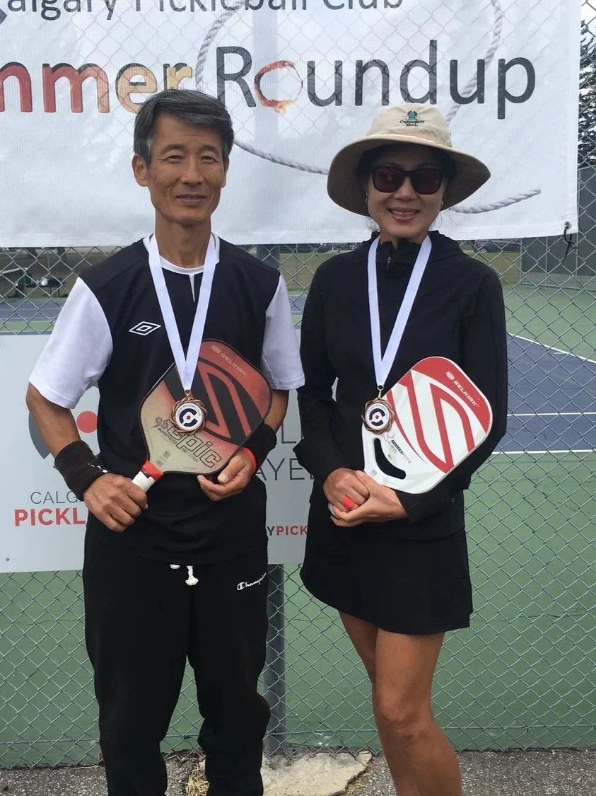 A man and woman standing in front of a fence at a pickleball tournament, holding pickleball paddles and wearing medals around their necks.