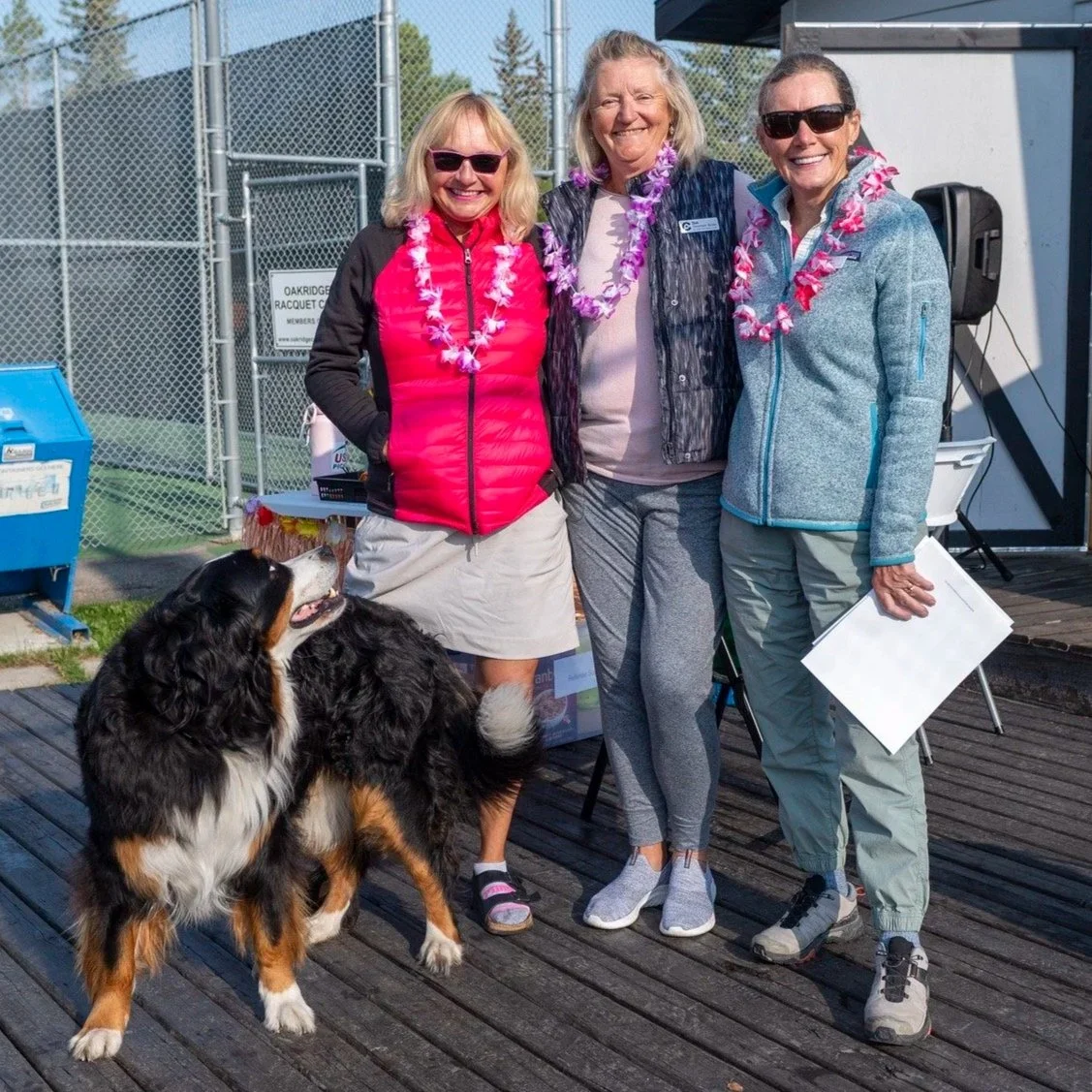 Three women standing outdoors on a wooden deck, smiling and wearing leis, with a large Bernese Mountain Dog. One woman holds a paper, and they are near a chain-link fence and sports field, with a blue donation box nearby.