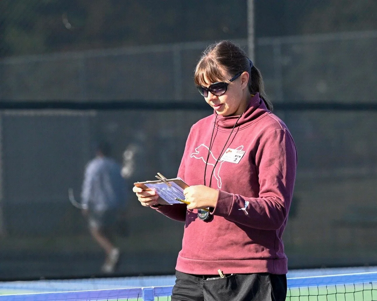 A woman wearing a maroon Puma hoodie and sunglasses stands outdoors near a tennis court, holding a clipboard or notepad.