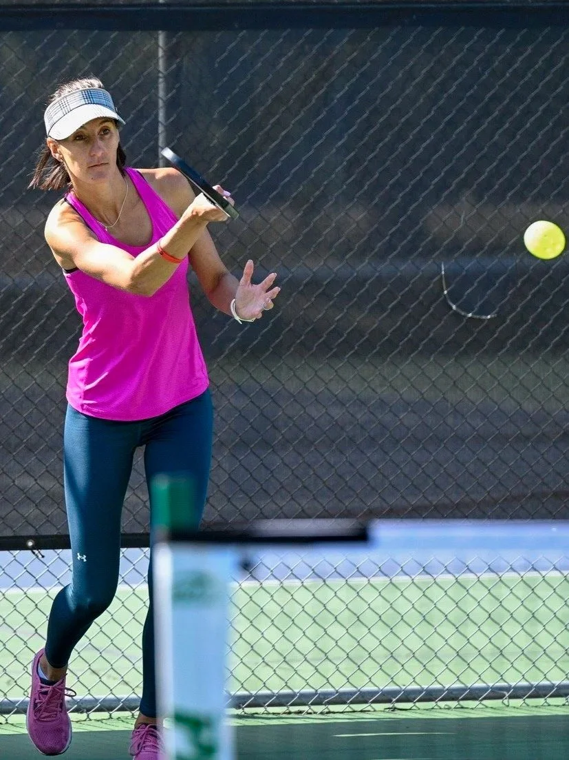 A woman in a pink tank top, black leggings, and a white visor playing tennis on an outdoor court, hitting a tennis ball with a racket.
