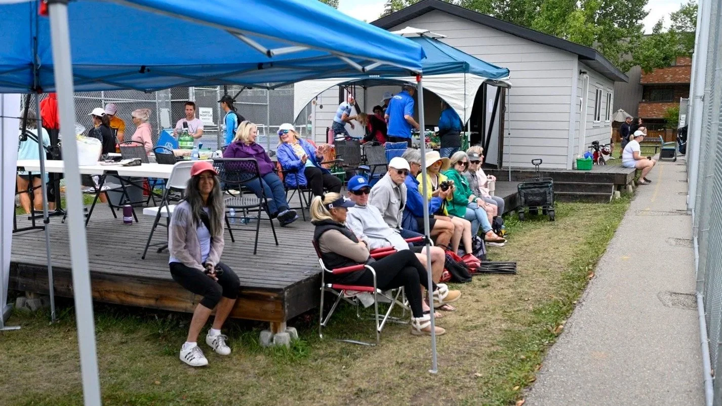People sitting outdoors on a wooden deck, some under blue canopies, at a social gathering or event during the daytime.