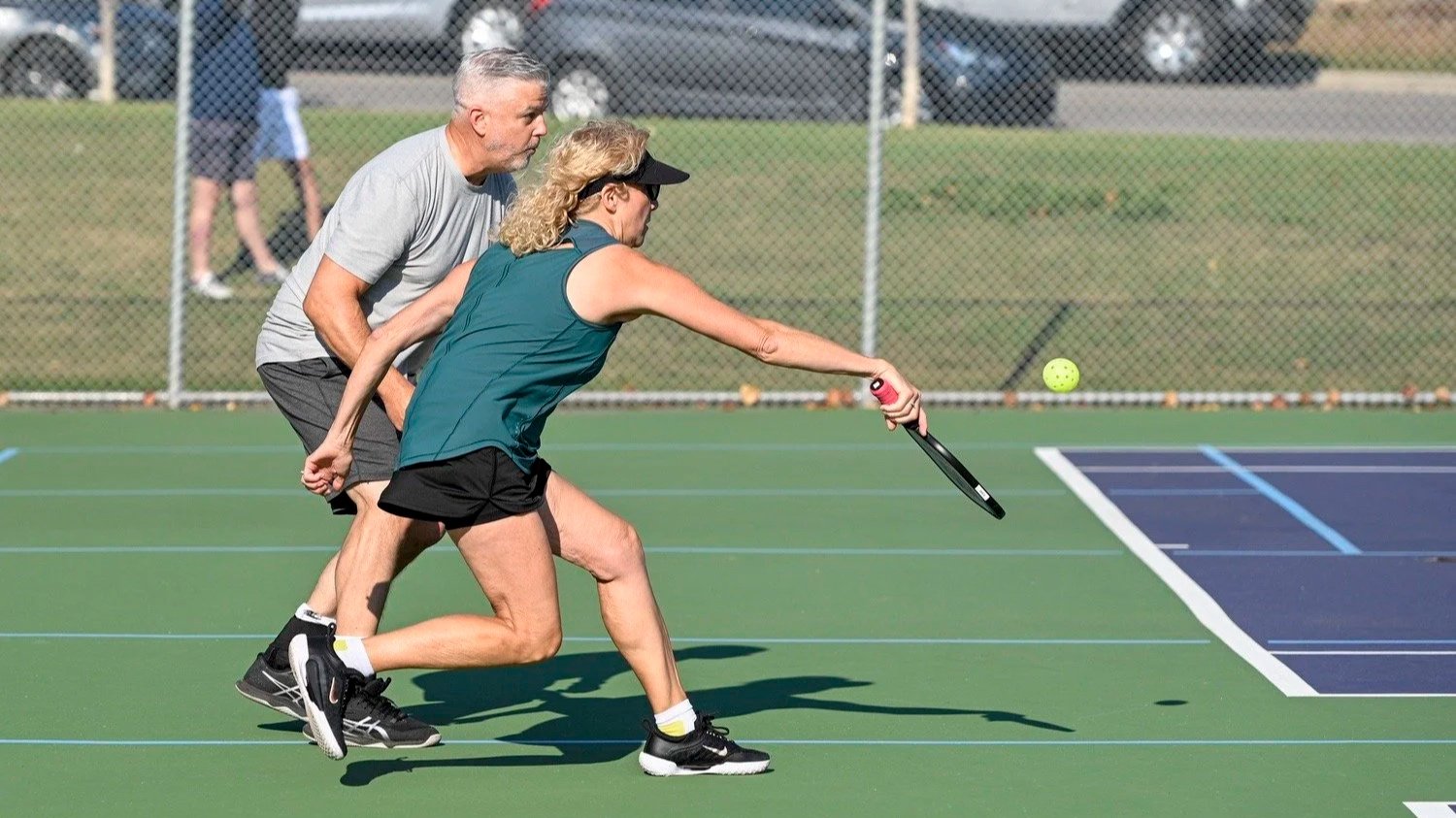 Two adults playing pickleball outdoors on a green court, with a chain-link fence and parked cars in the background.