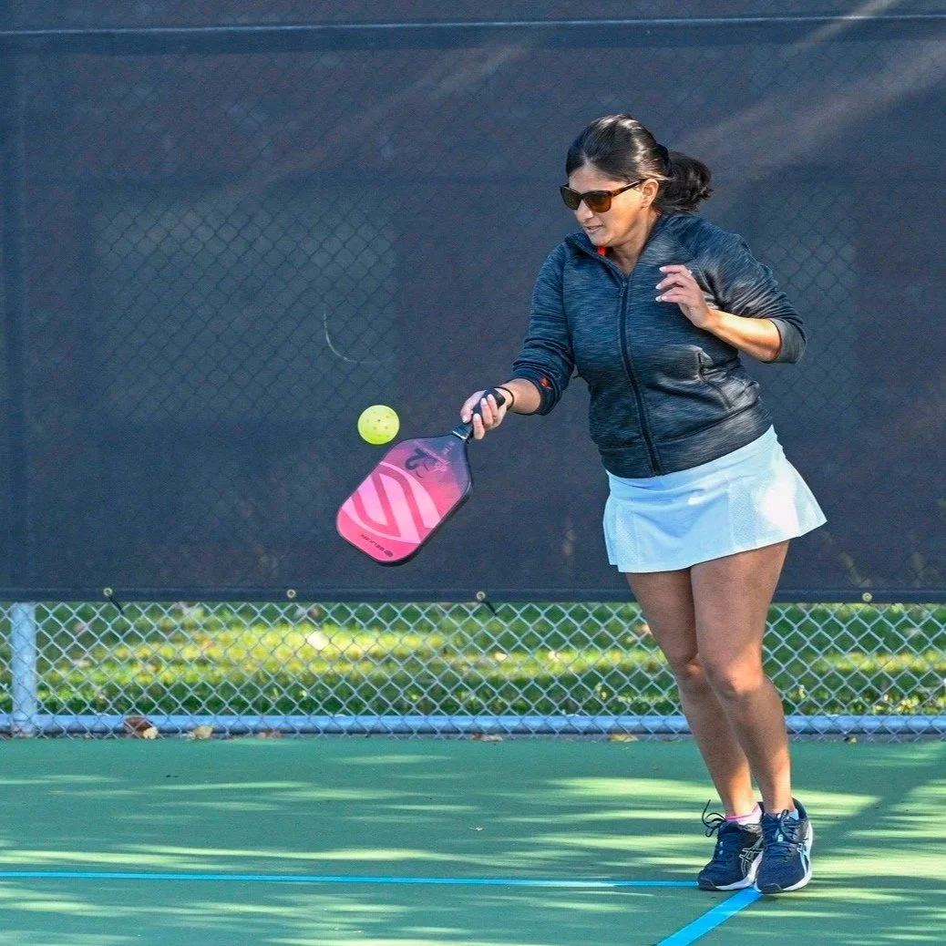 A woman in sunglasses, a dark jacket, and a white skirt playing pickleball on an outdoor court, hitting a yellow ball with a pink paddle.
