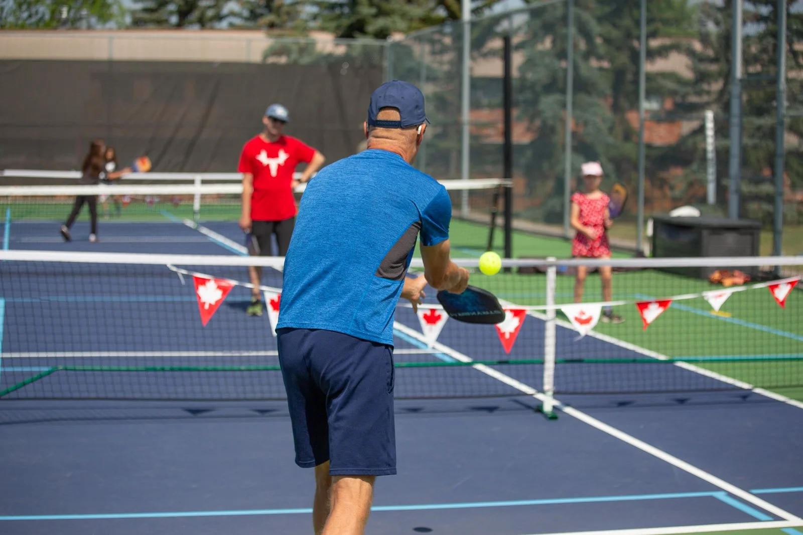 Adults and children playing pickleball on an outdoor court decorated with a Canadian flag banner, with trees and a fence in the background.