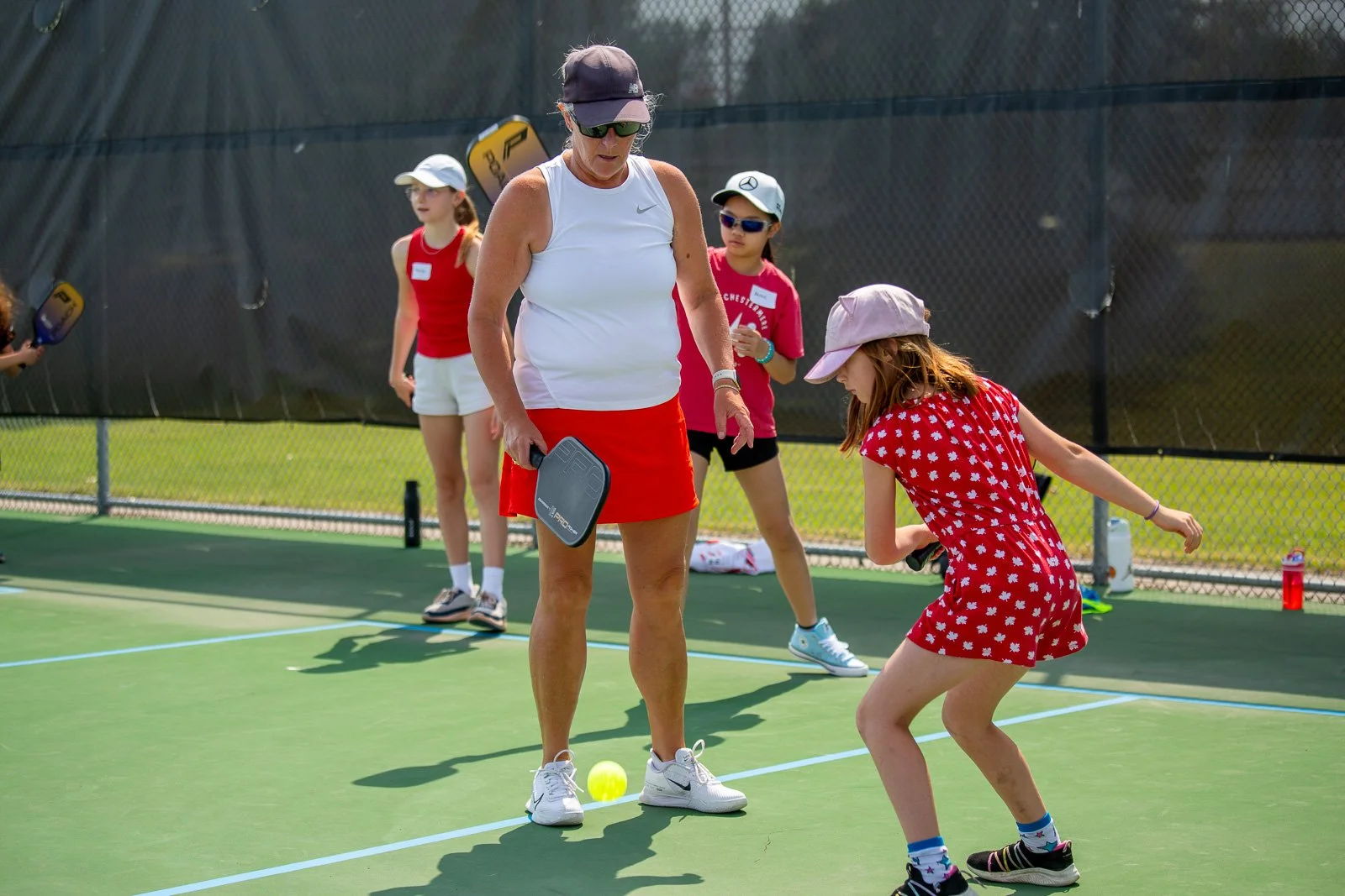 A woman teaching children how to play tennis on a tennis court, with a young girl in a red dress in the foreground, holding a tennis racket, and other children watching in the background.