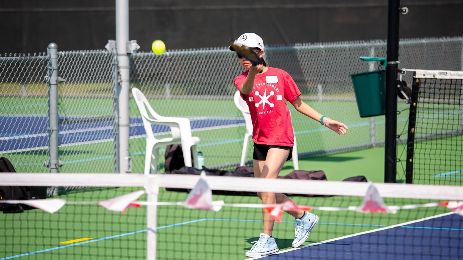A young girl playing tennis on a court, wearing a white cap, red shirt, black shorts, and white sneakers