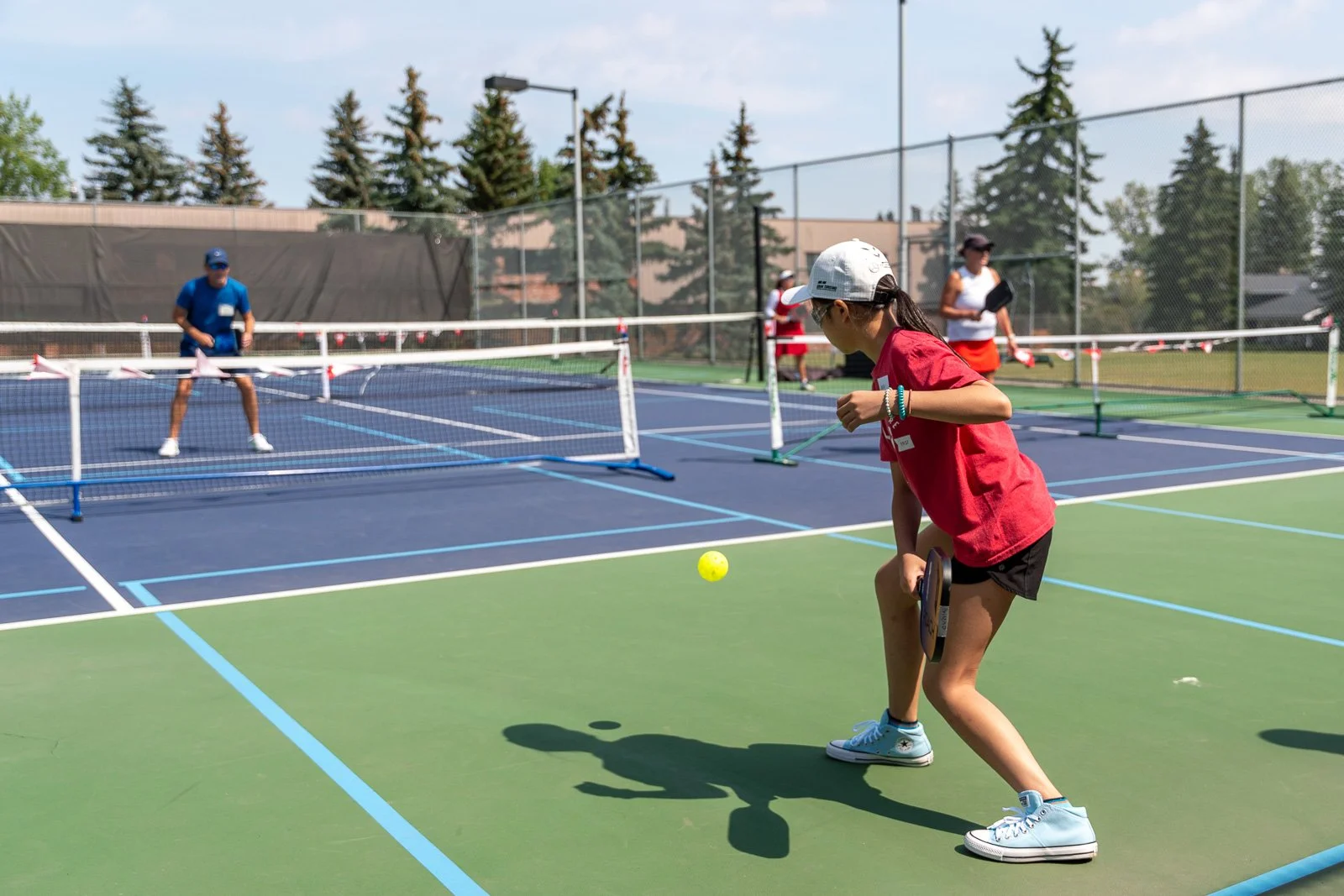 Kids playing pickleball on an outdoor courts, with a girl in a red shirt and white cap preparing to hit a yellow ball, and a boy in a blue shirt and cap standing near the net.