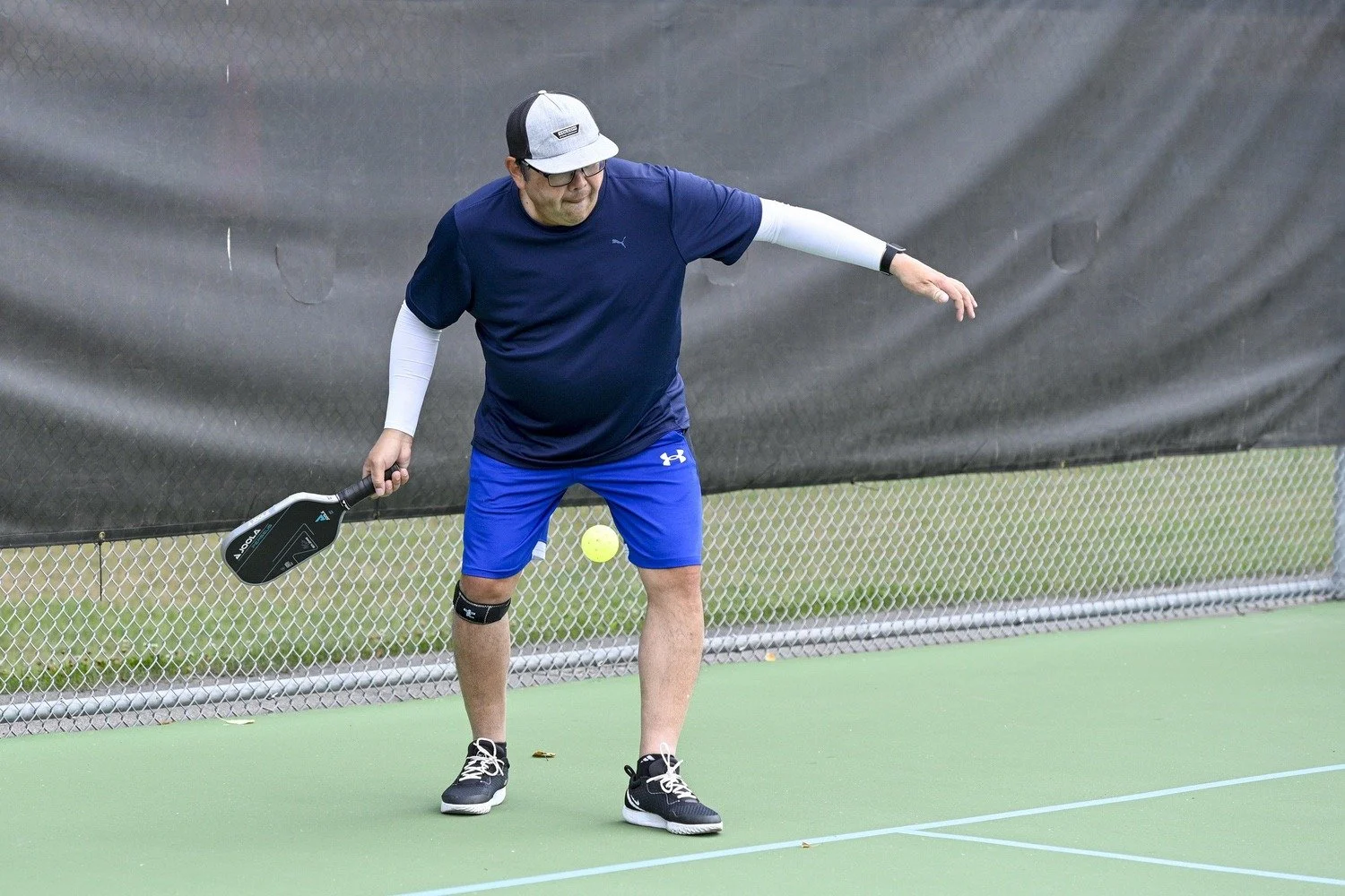 A man wearing a navy blue t-shirt, blue shorts, and black sneakers playing pickleball on a green court. He is holding a black pickleball paddle in his right hand and preparing to hit a yellow pickleball.