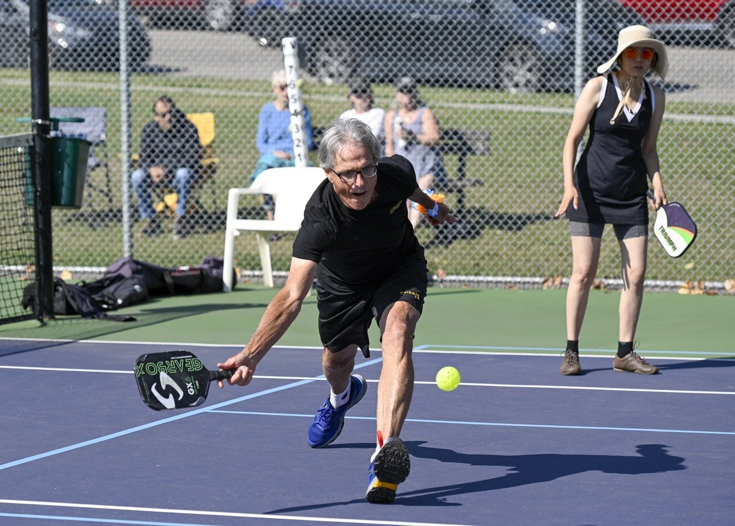 An elderly man playing pickleball on a tennis court. He is crouched down, reaching for a bright green ball with a paddle in his right hand. A woman stands nearby holding a paddle, wearing sunglasses, a large sunhat, and casual clothing. There are spe