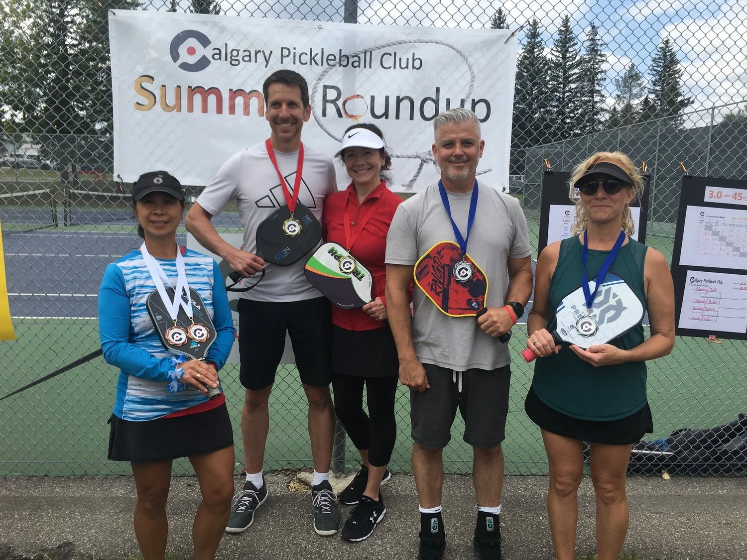 Group of five people standing in front of a chain-link fence with a banner that reads 'Calgary Pickleball Club Summer Roundup'. The individuals are smiling, holding paddle rackets, and wearing medals around their necks, indicating they participated i