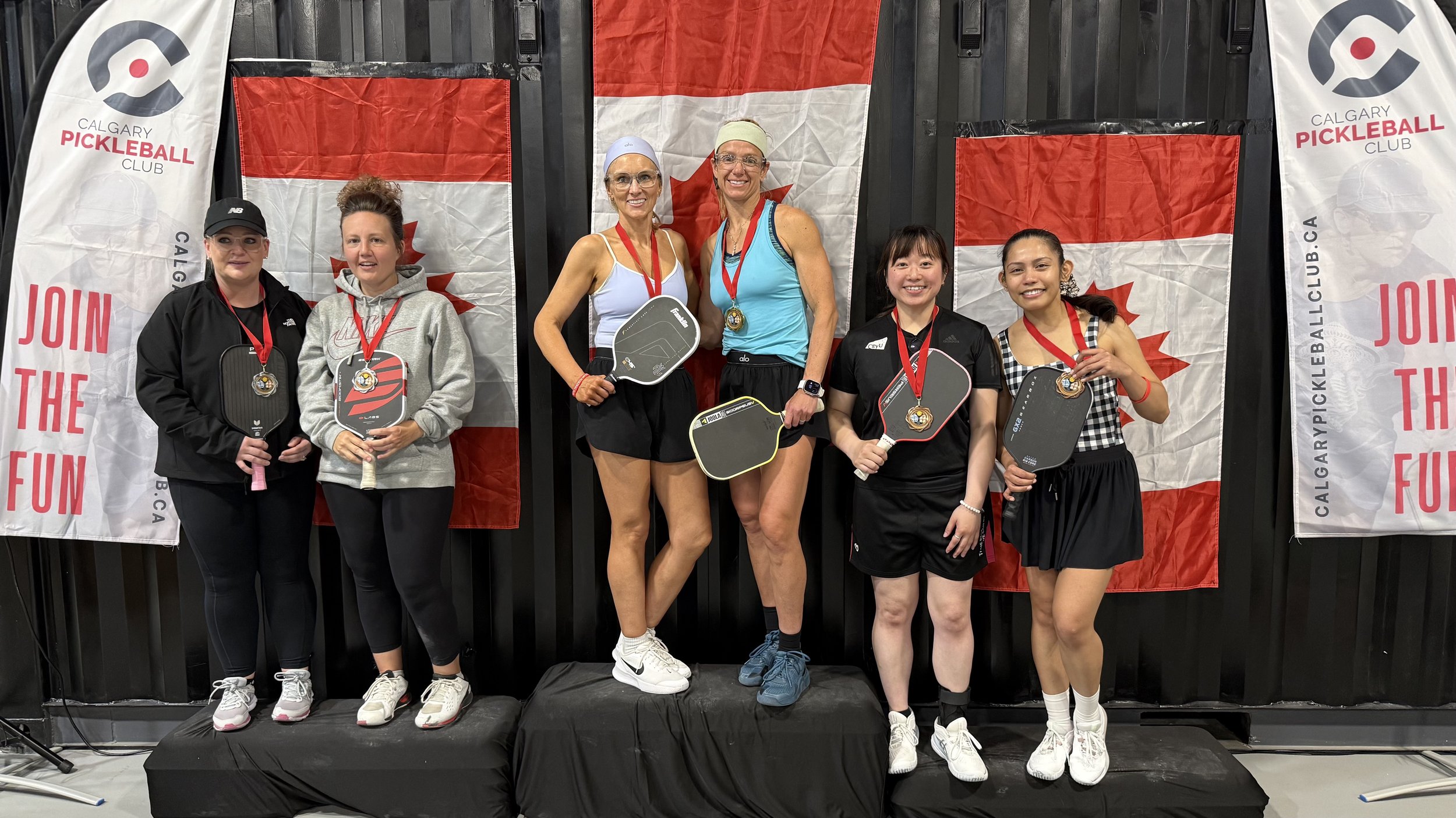 Group of six women standing on a podium, holding pickleball paddles and wearing medals, in front of Calgary Pickleball Club banners and flags.