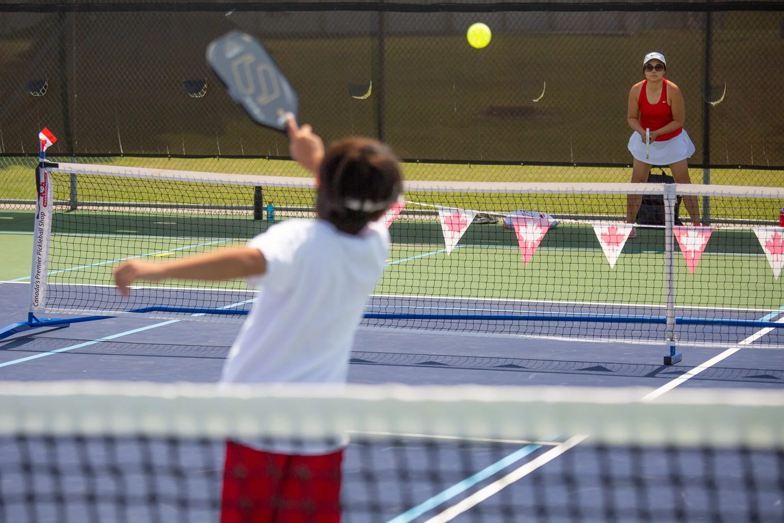 A boy and a woman playing pickleball on an outdoor court with a tennis net and pink bunting