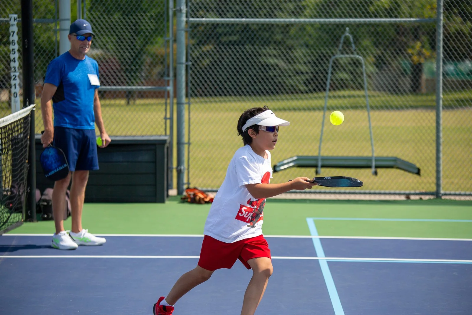 A young boy playing tennis on an outdoor court, wearing a white cap, sunglasses, a white T-shirt, red shorts, and holding a tennis racket, with an adult man watching nearby.