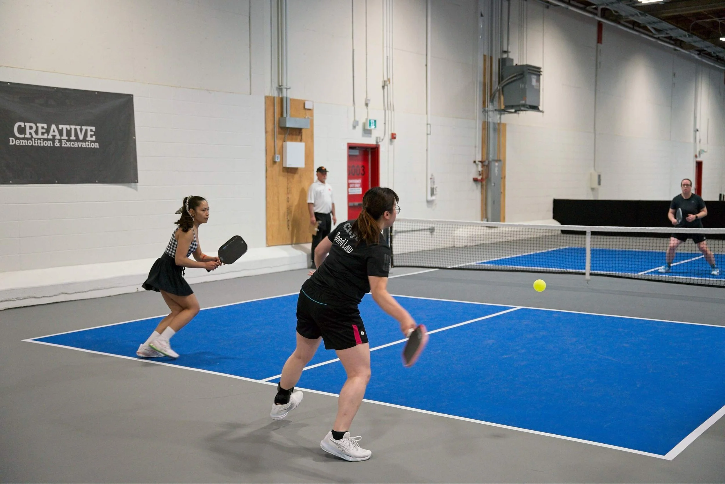 People playing pickleball indoors on a blue court, with a net in the middle, and a large sign on the wall that reads 'CREATIVE Demolition & Excavation'.