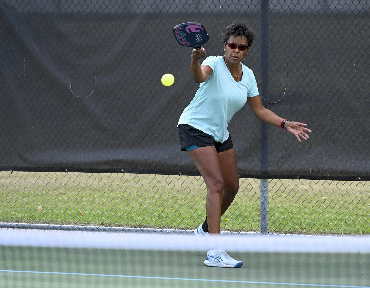 Woman playing pickleball on a court, wearing sunglasses, a light blue shirt, black shorts, and holding a paddle, about to hit a yellow pickleball.