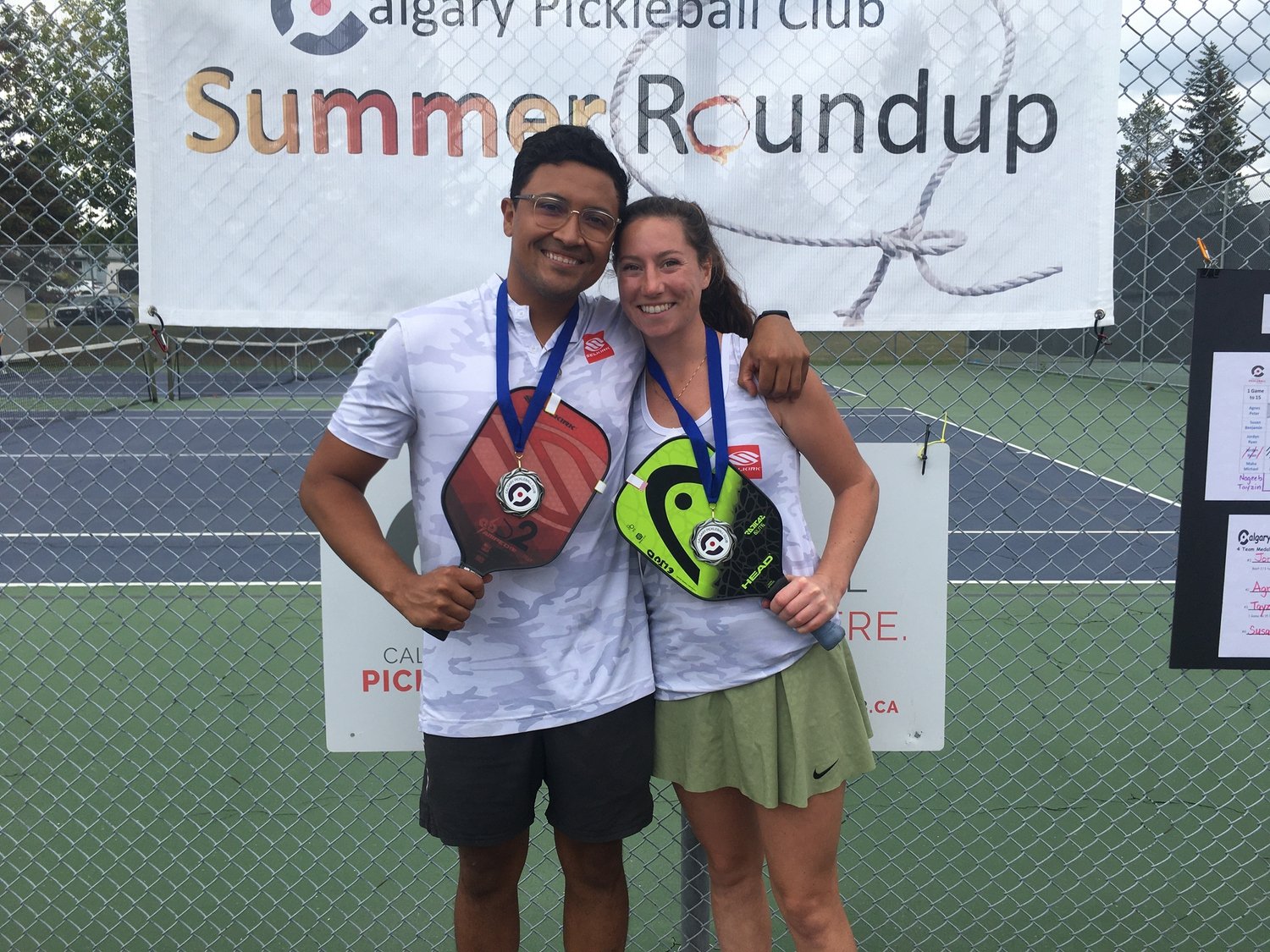 Two people, a man and a woman, standing side by side on a tennis court with a chain-link fence behind them. They are smiling and holding tennis rackets, wearing medals around their necks. A large banner above them reads 'Calgary Pickleball Club Summe