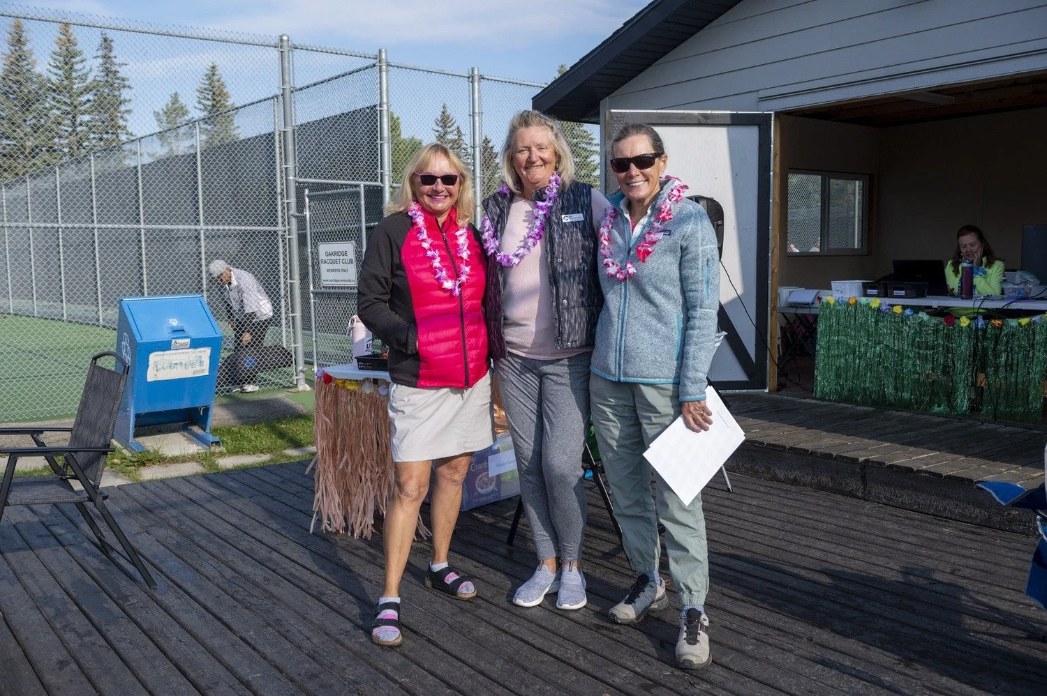 Three women with leis around their necks standing on a wooden deck outdoors, smiling at the camera. Two of them are wearing sunglasses. One woman is holding a piece of paper. In the background, there is a tennis court with a chain-link fence, and ano