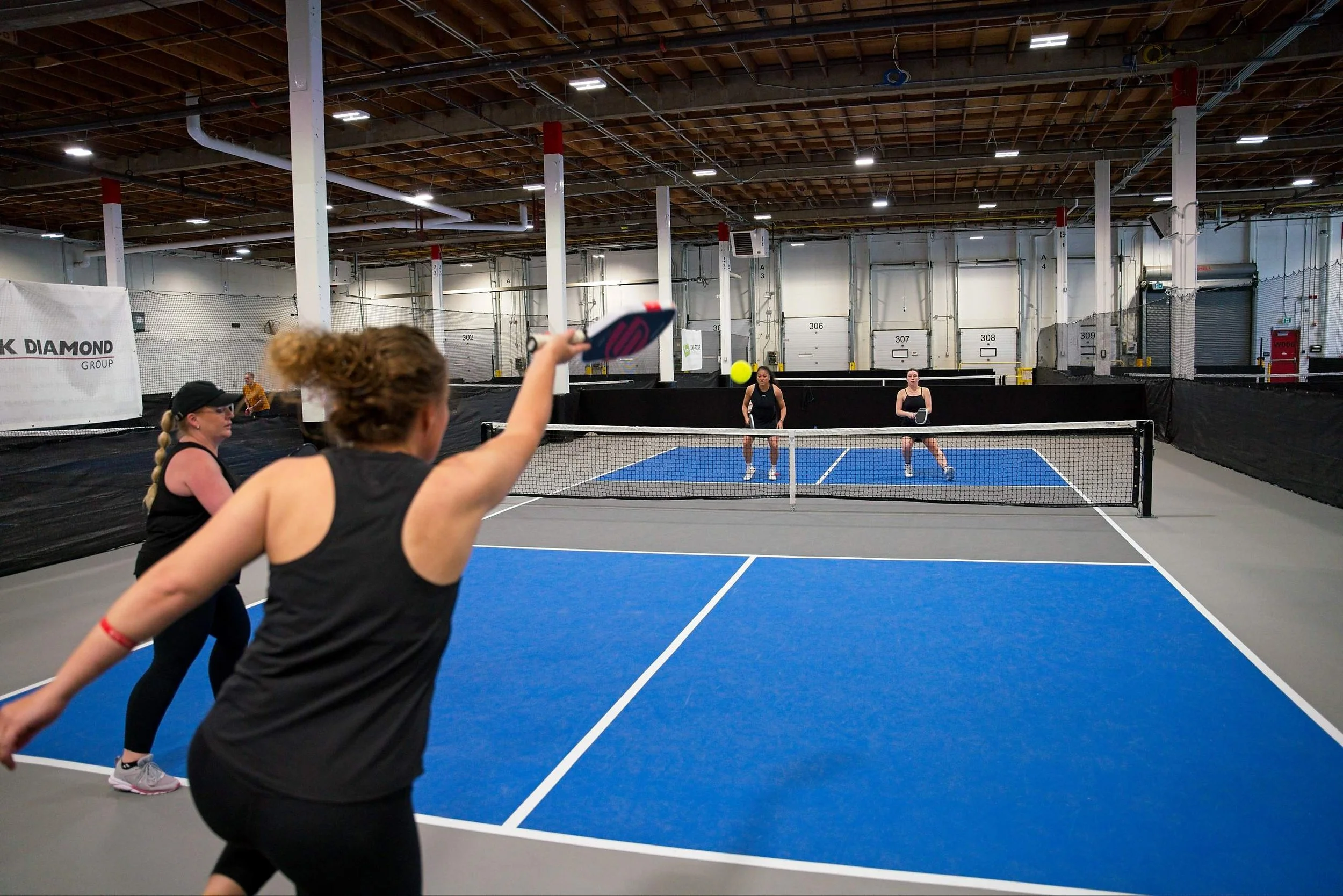 People playing pickleball indoors on a blue court with white lines, hitting a yellow ball with paddles.