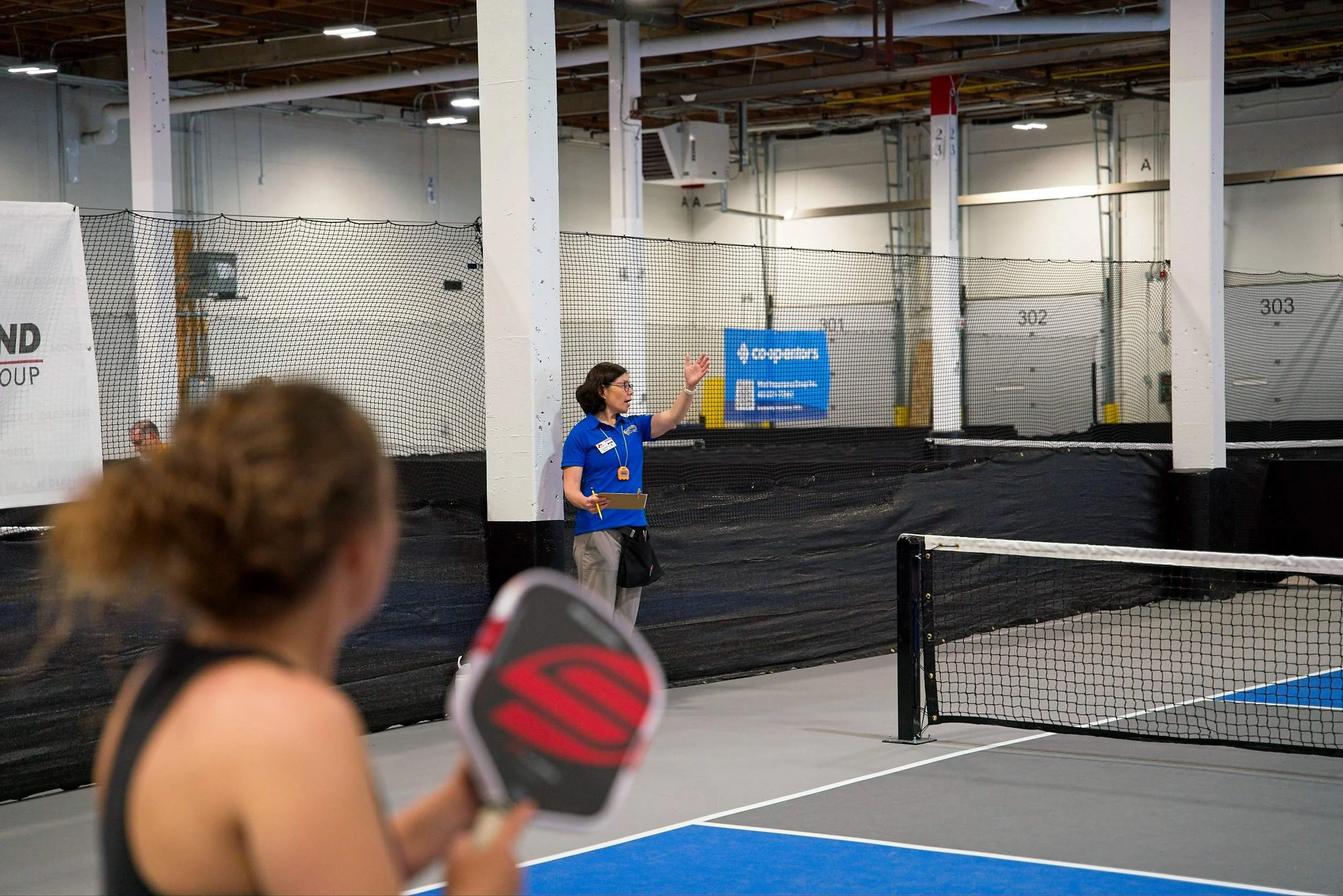 A woman in a blue shirt is speaking and gesturing in front of a tennis court with a net, with a person holding a tennis paddle in the foreground. The background shows a large indoor sports facility with high ceilings and numbered storage units.