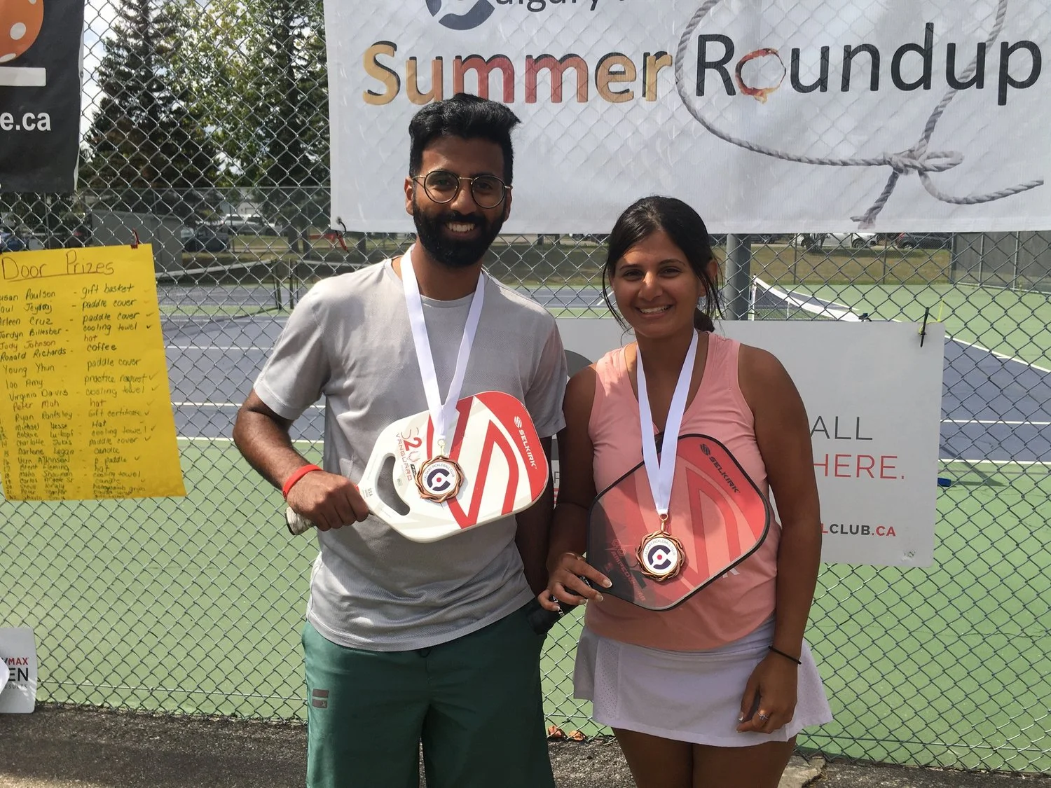 A man and a woman standing outdoors on a tennis court, smiling and holding medals and paddle covers, with a banner and a yellow list of names in the background.
