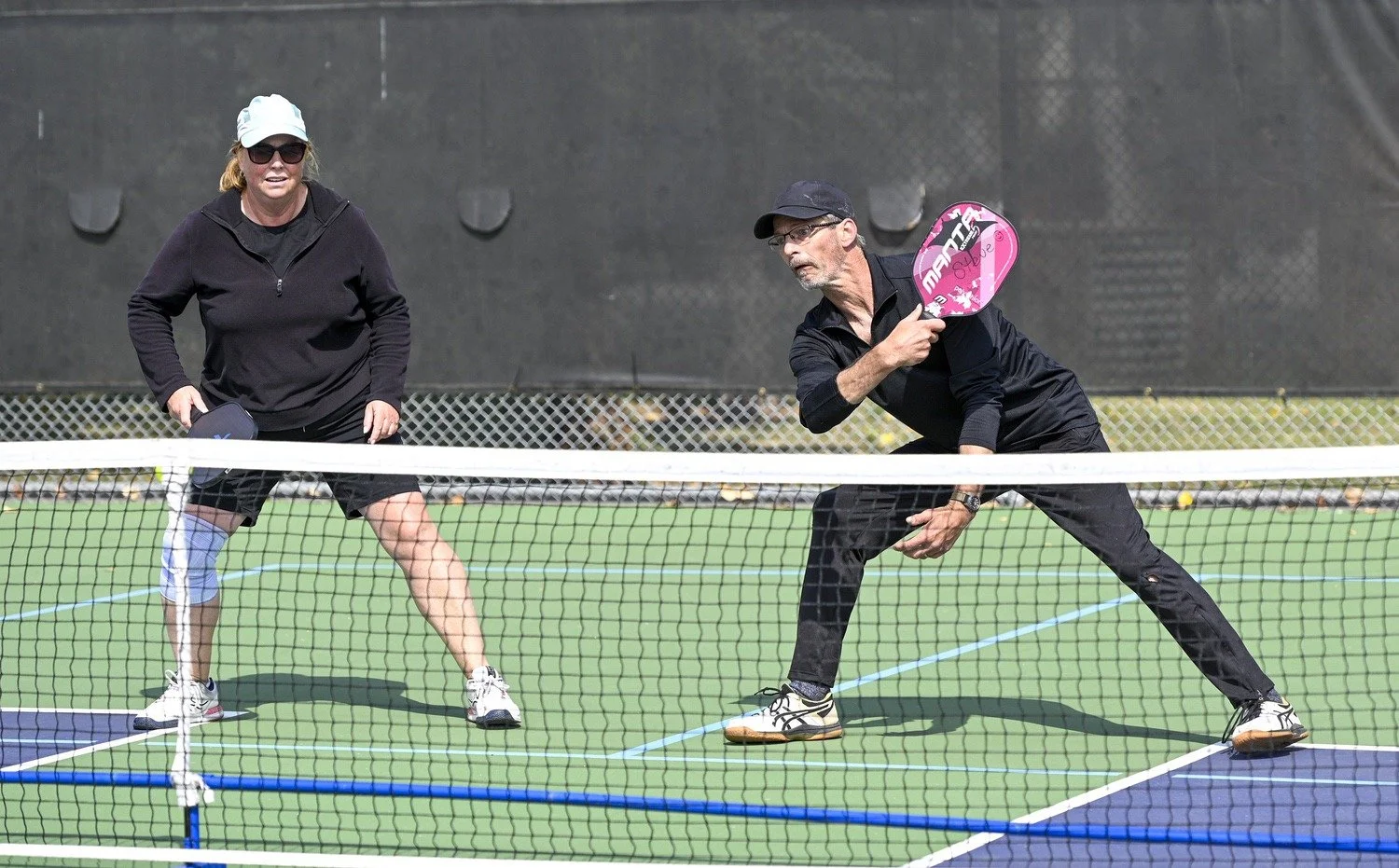 Two people playing pickleball on an outdoor court. The woman on the left is wearing sunglasses, a white cap, a black jacket, and shorts. The man on the right is wearing glasses, a black cap, a black long-sleeve shirt, and black pants. He is holding a