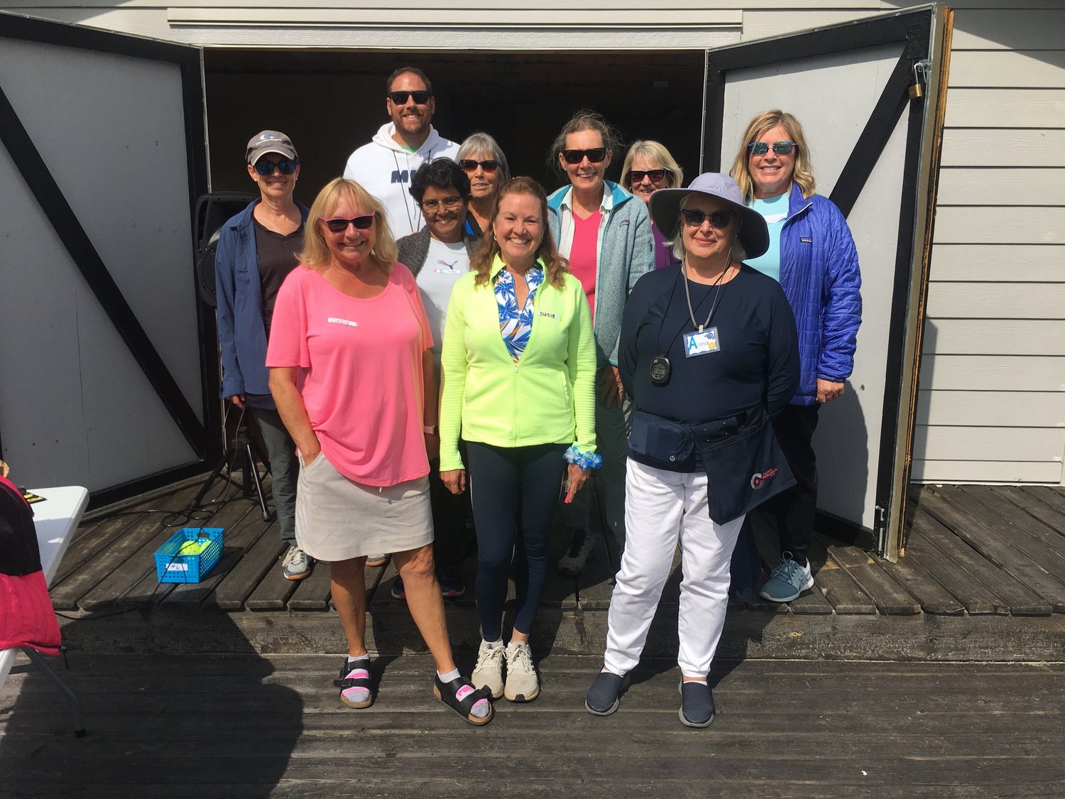 Group of eleven smiling people standing on a wooden deck outside a shed, posing for a photo on a sunny day.