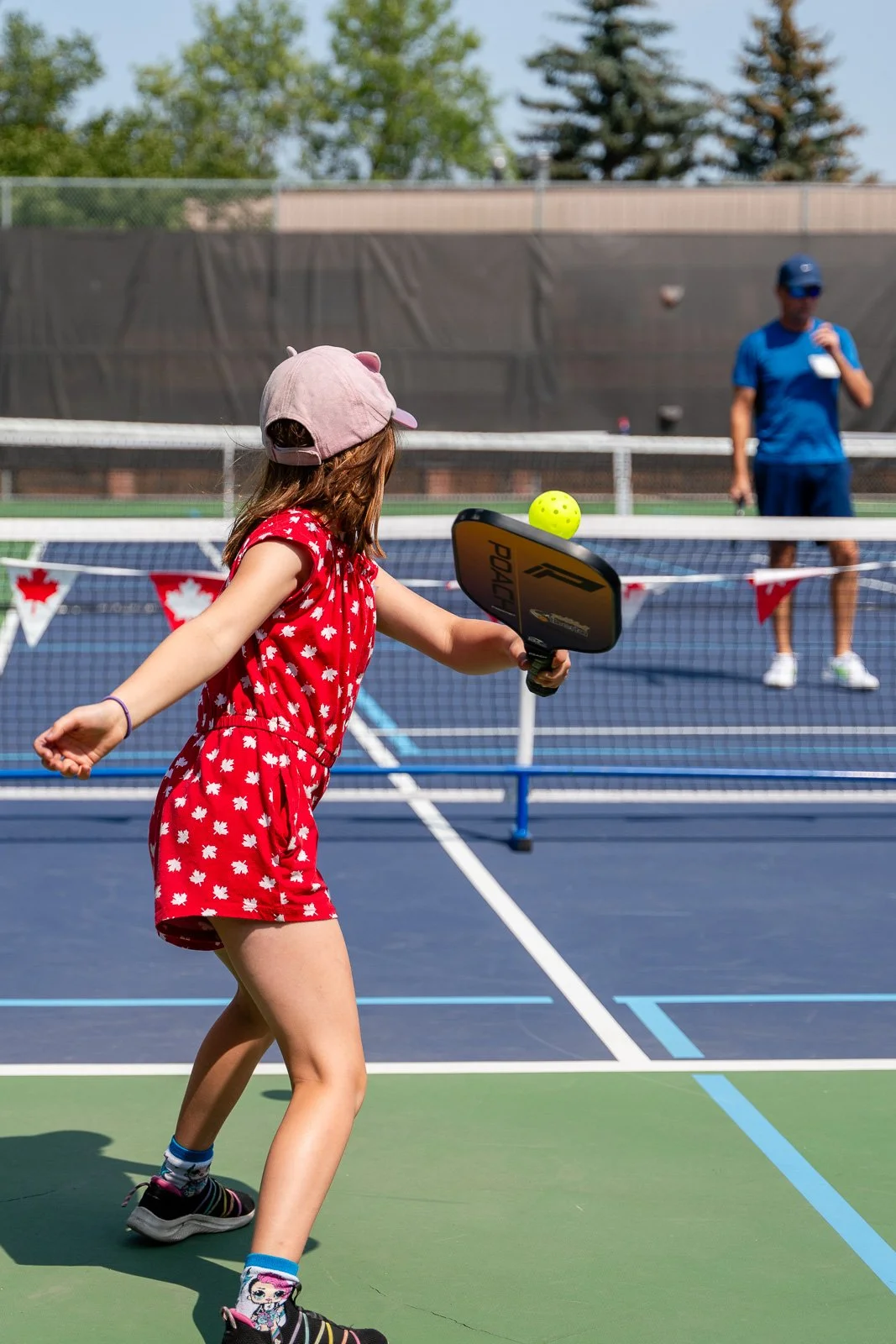 A young girl in a red dress and pink cap playing pickleball on an outdoor court, with a man in a blue shirt and cap in the background.